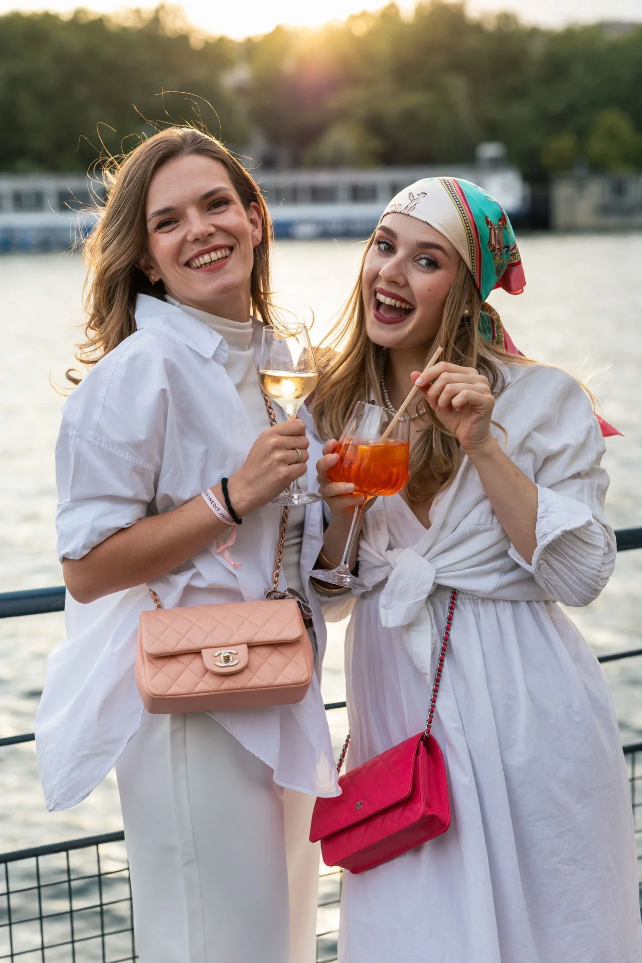 Deux femmes souriantes posent avec des boissons au bord d'une rivière, l'une tenant un verre de vin blanc et l'autre un cocktail orangé; l'une porte un bandana coloré et une petite sacoche rose, l'autre une veste blanche et une sacoche beige.