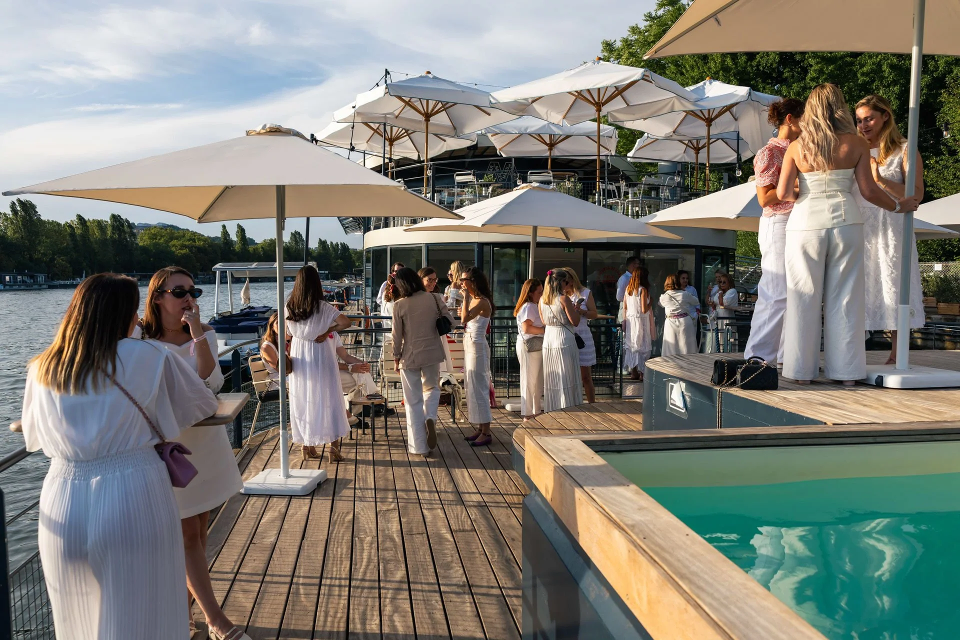 Groupe de personnes sur un bateau-restaurant au bord de l'eau avec parasols blancs, certains discutant et d'autres regardant vers le pont supérieur, dans un cadre verdoyant par une belle journée.