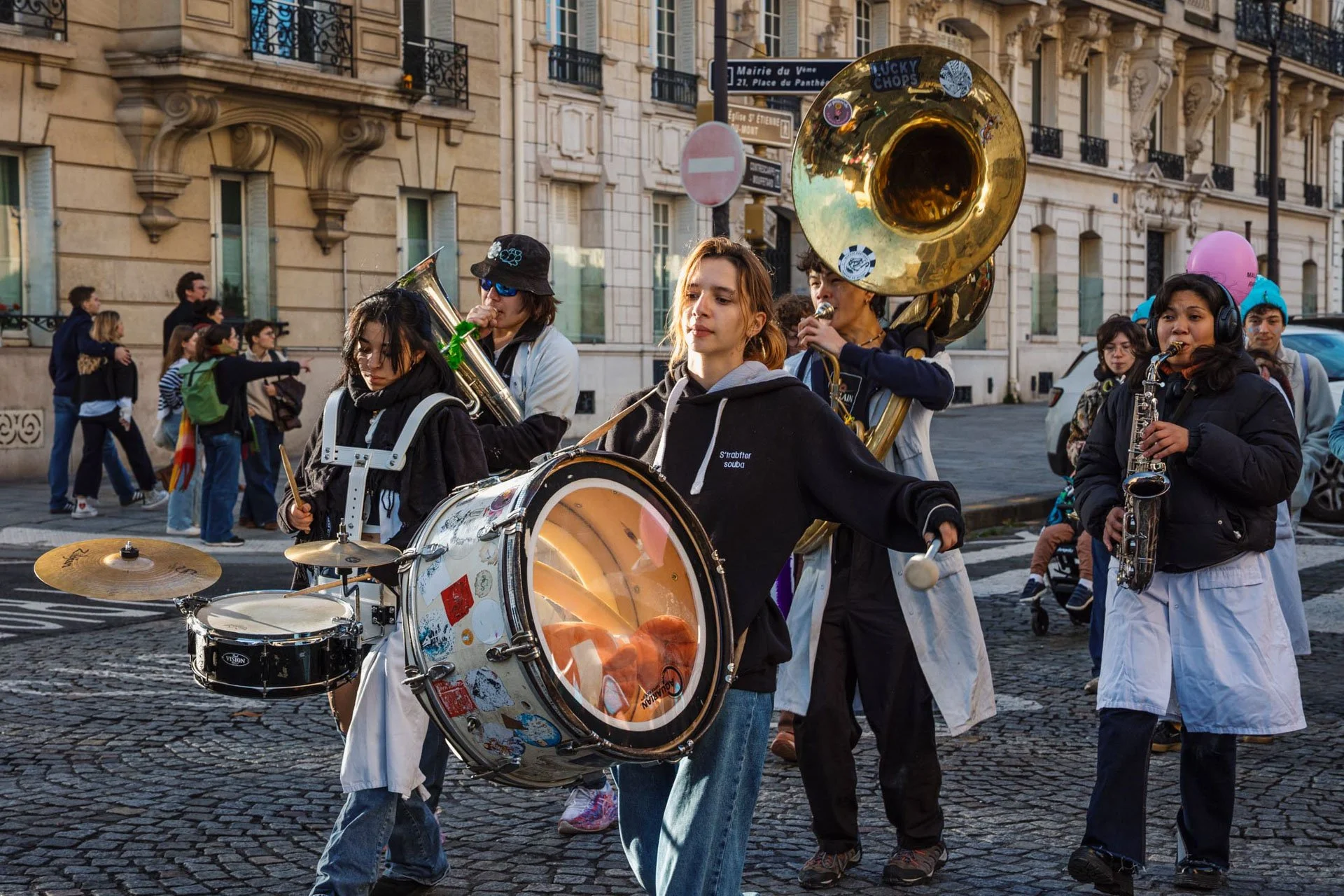 Un groupe de jeunes musiciens joue dans une parade urbaine, avec un bâtiment historique en arrière-plan, lors d'une journée ensoleillée.