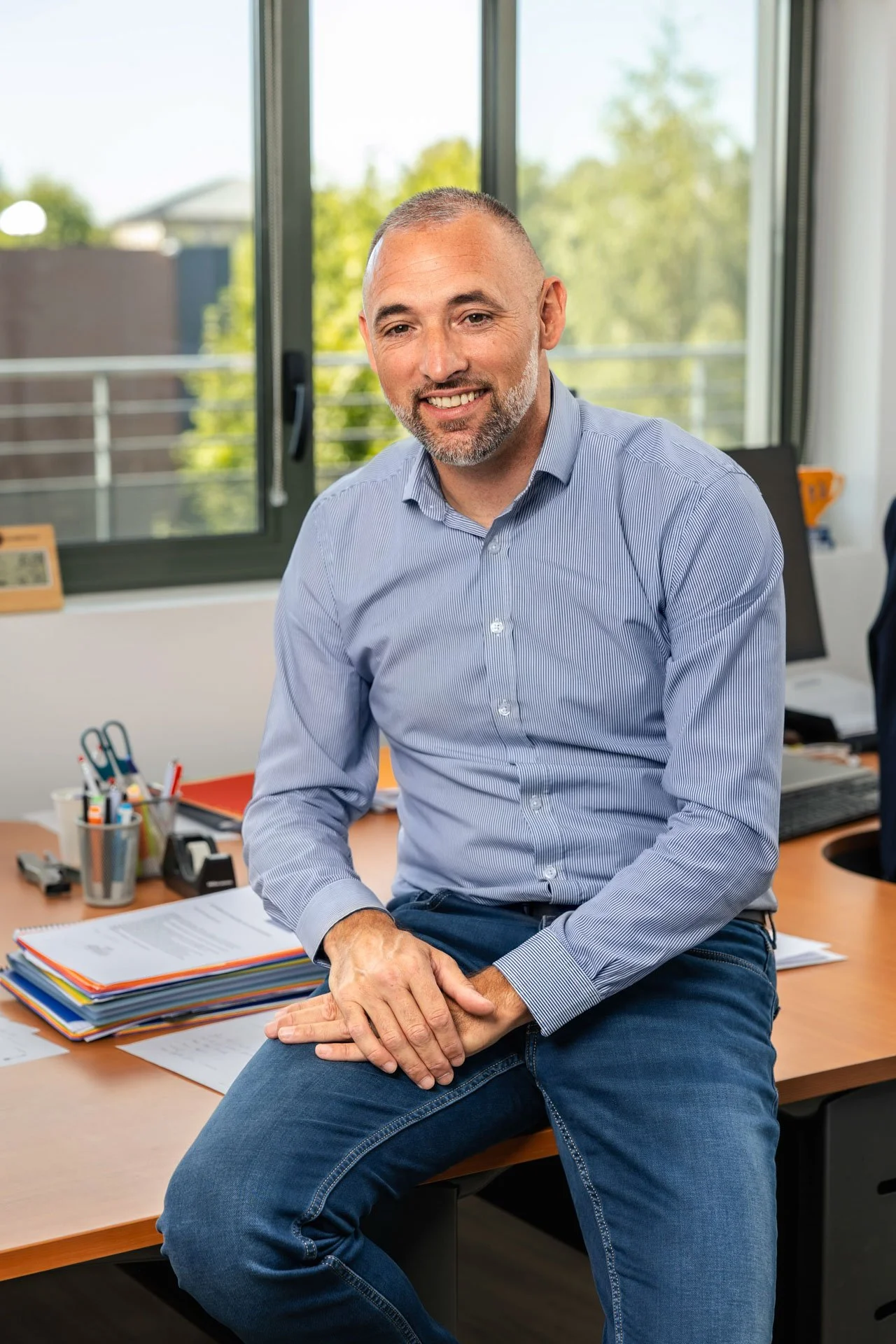 Un homme homme assis sur un bureau en milieu de travail, portant une chemise bleue et un pantalon en jean, avec un environnement de bureau en arrière-plan.