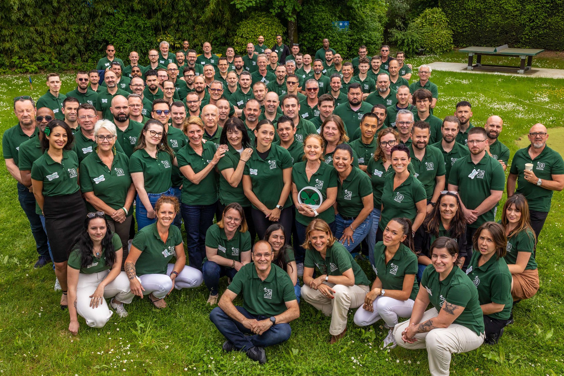 Photo de groupe de plusieurs personnes portant des polos verts, posant à l'extérieur sur une pelouse entourée d'arbres, lors d'une célébration ou d'un événement.