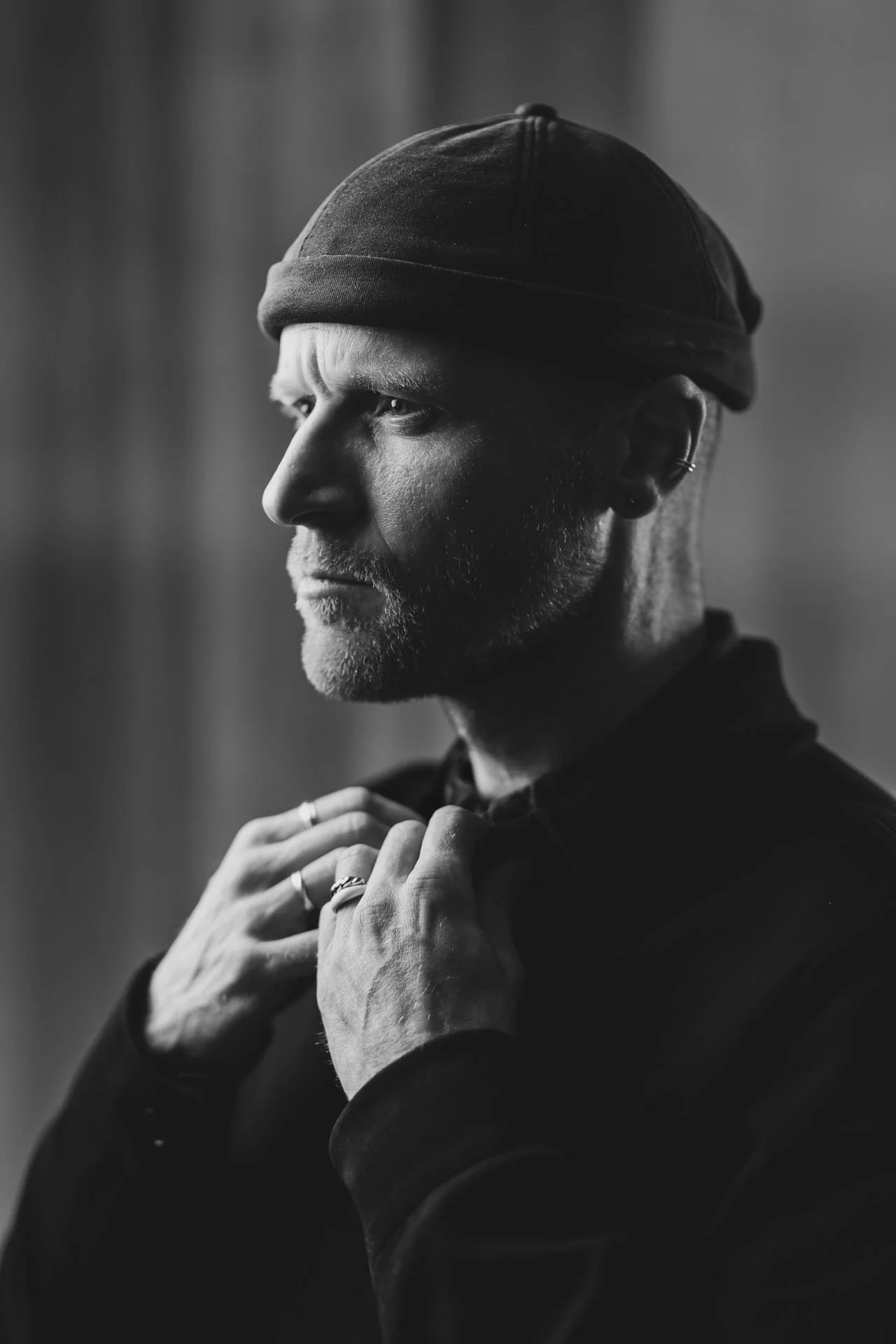 Portrait en noir et blanc d'un homme portant un bonnet, regardant vers la gauche, avec un regard concentré, mains près du col, plusieurs rings visibles à ses doigts.