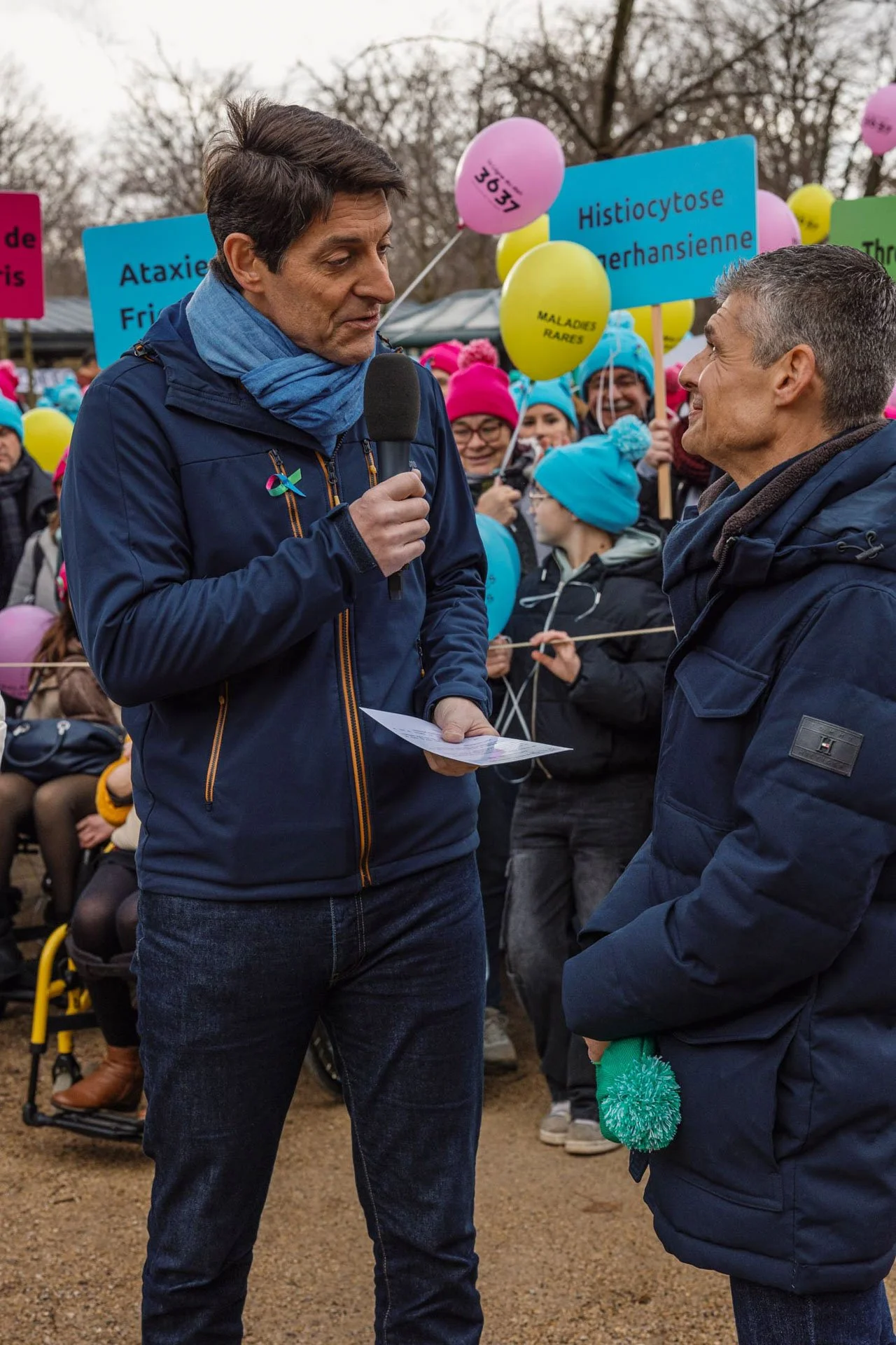 Deux hommes se parlent lors d'une manifestation de soutien à la recherche sur la neurofibromatose, entourés de manifestants avec des ballons colorés portant des messages en français.