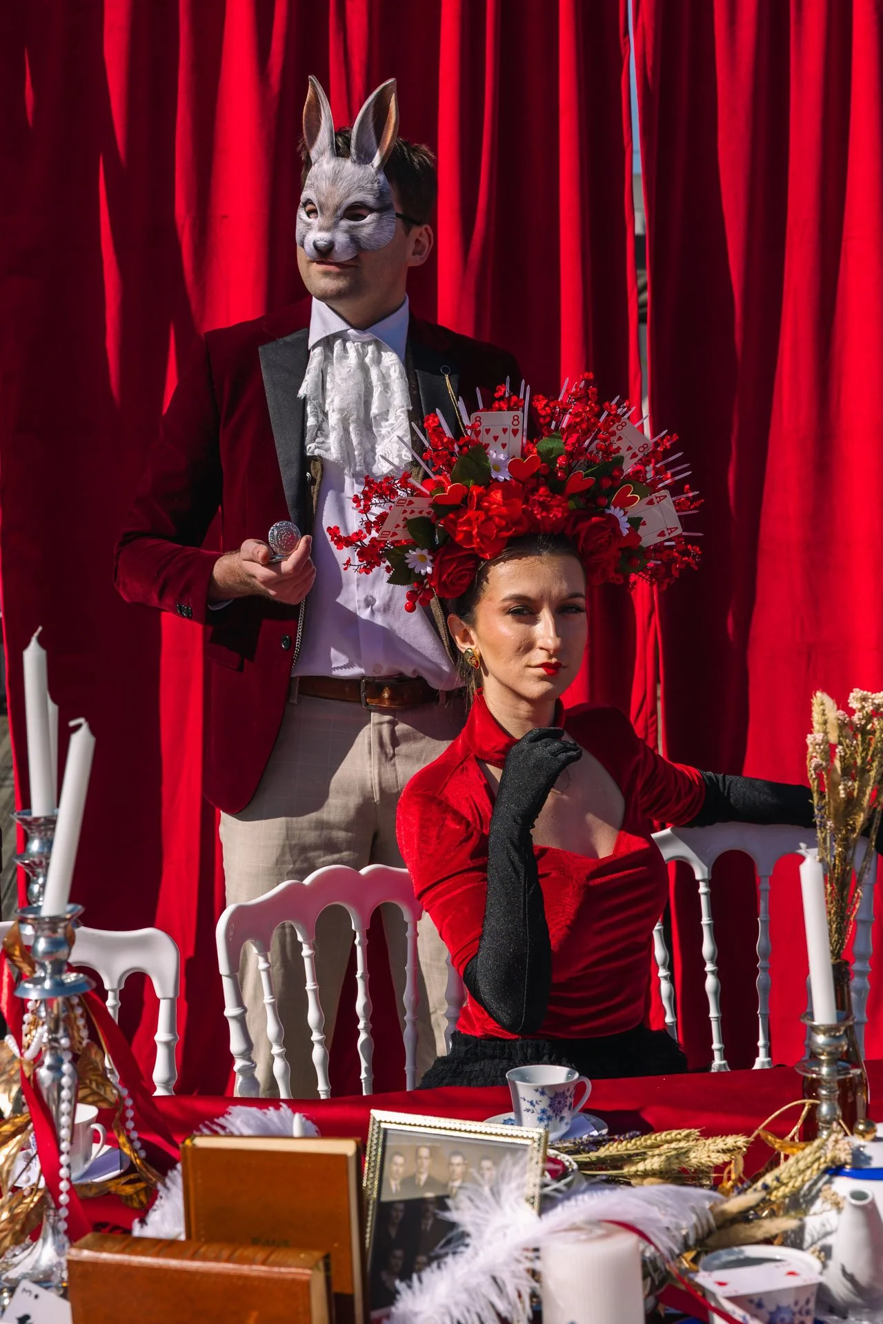 Une scène théâtrale avec deux personnes, une avec un masque de lapin et une autre avec une couronne de fleurs rouges, dans un décor avec des rideaux rouges.