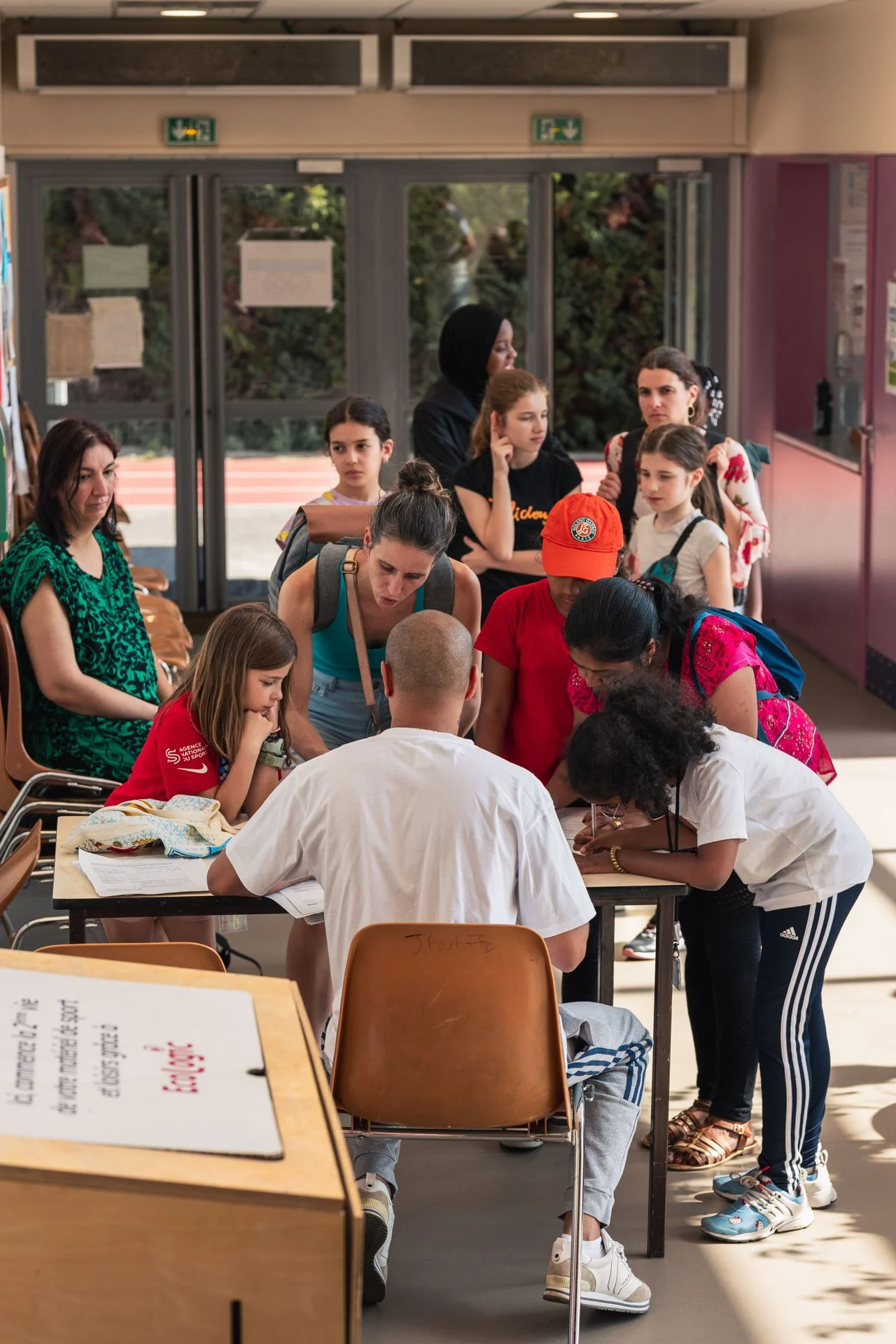 Groupe de personnes, majoritairement des jeunes, attendant devant une table avec deux personnes assises, probablement pour s'inscrire ou obtenir des informations dans un lieu intérieur.