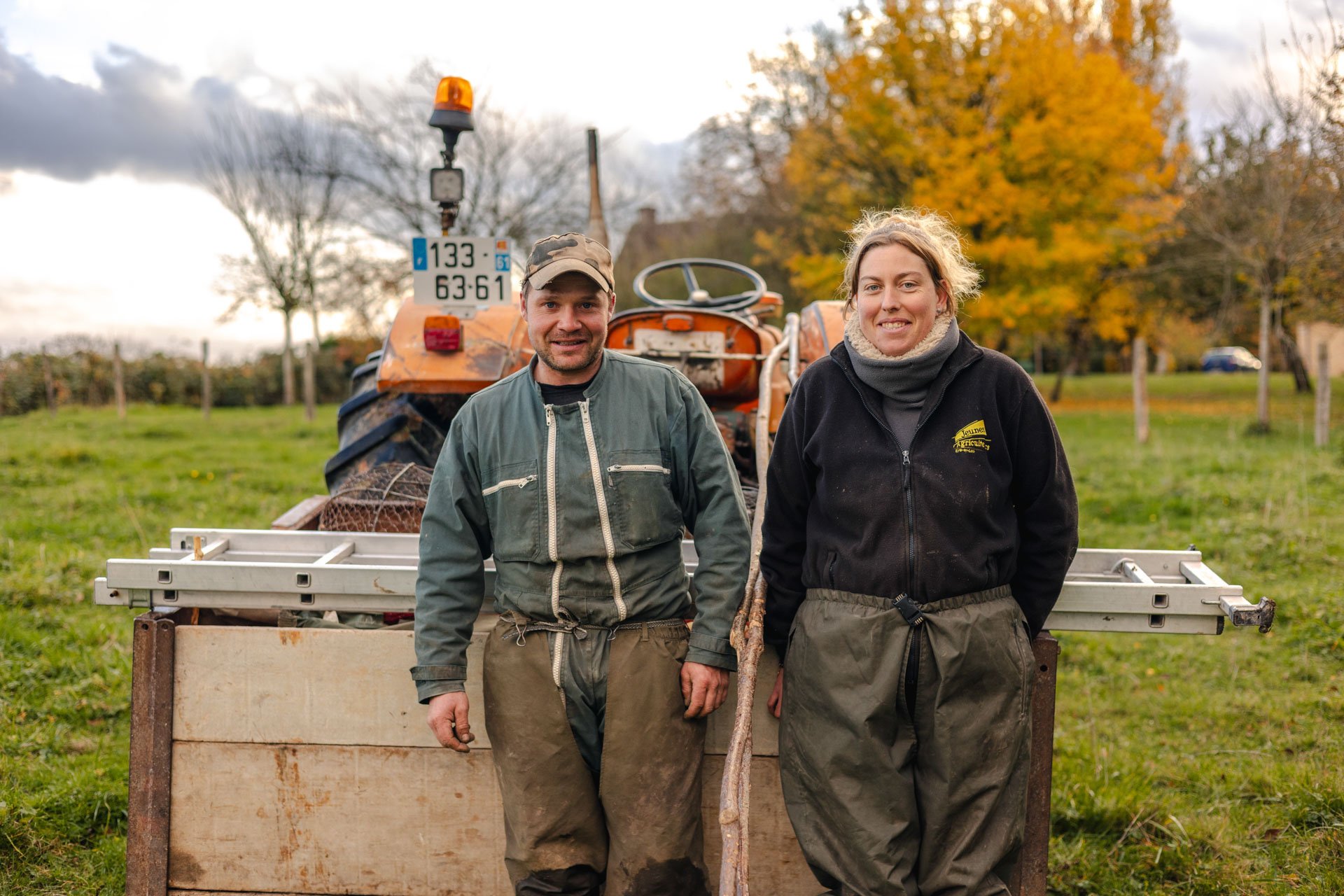 Deux personnes posent devant un tracteur orange dans un champ avec des arbres aux feuilles automnales.