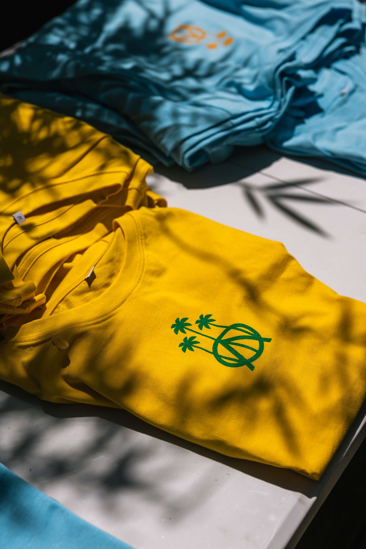 Un sweat jaune avec un logo vert de palmiers et un cercle, posé sur une surface blanche, avec des ombres de feuilles dessus. En arrière-plan, des vêtements bleus et gris sont visibles.