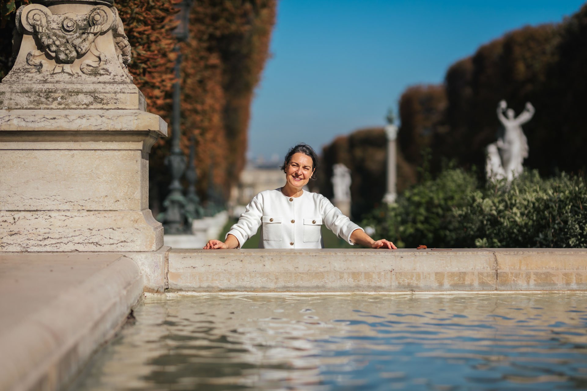 Une femme souriante en chemise blanche se tient derrière un muret en pierre au bord d'une fontaine, avec des statues classiques et des arbres feuillus en arrière-plan, par une journée ensoleillée.