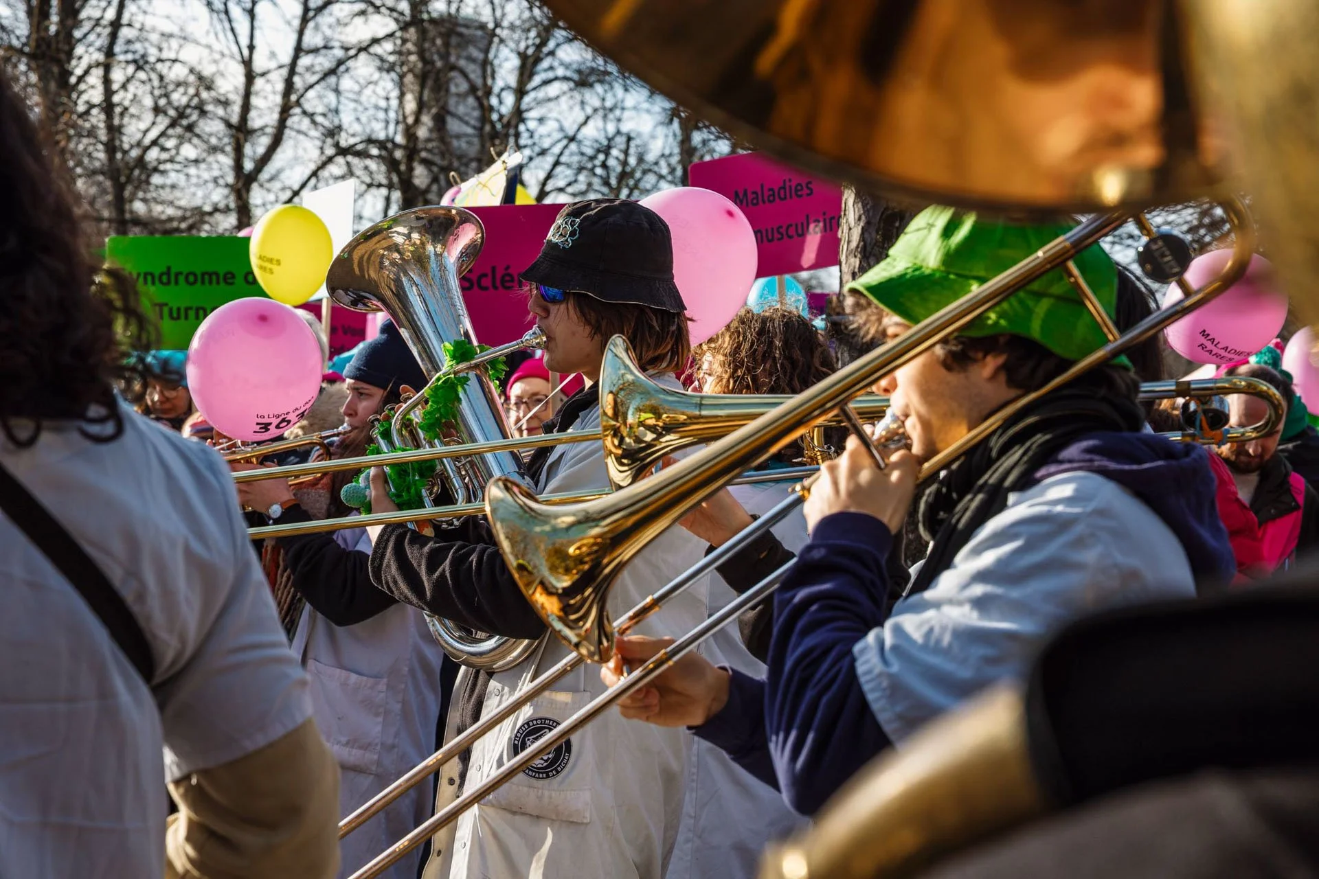 Groupe de musiciens jouant de la trompette lors d'une manifestation en plein air, avec des panneaux de protestation en arrière-plan, certains balloons et des arbres.