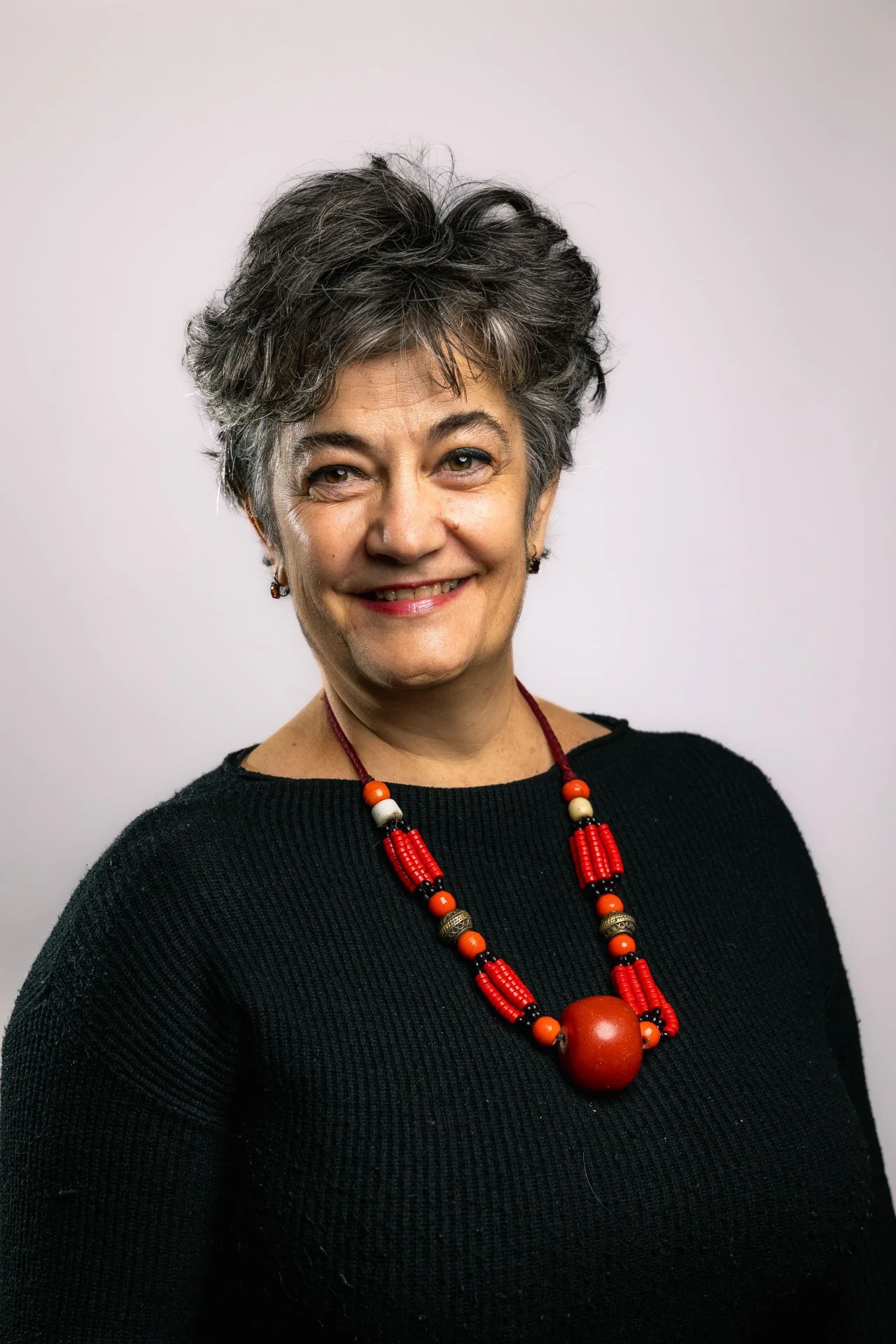 Portrait d'une femme d'âge moyen avec des cheveux courts gris et bouclés, portant un pull noir et un collier en perles rouges.