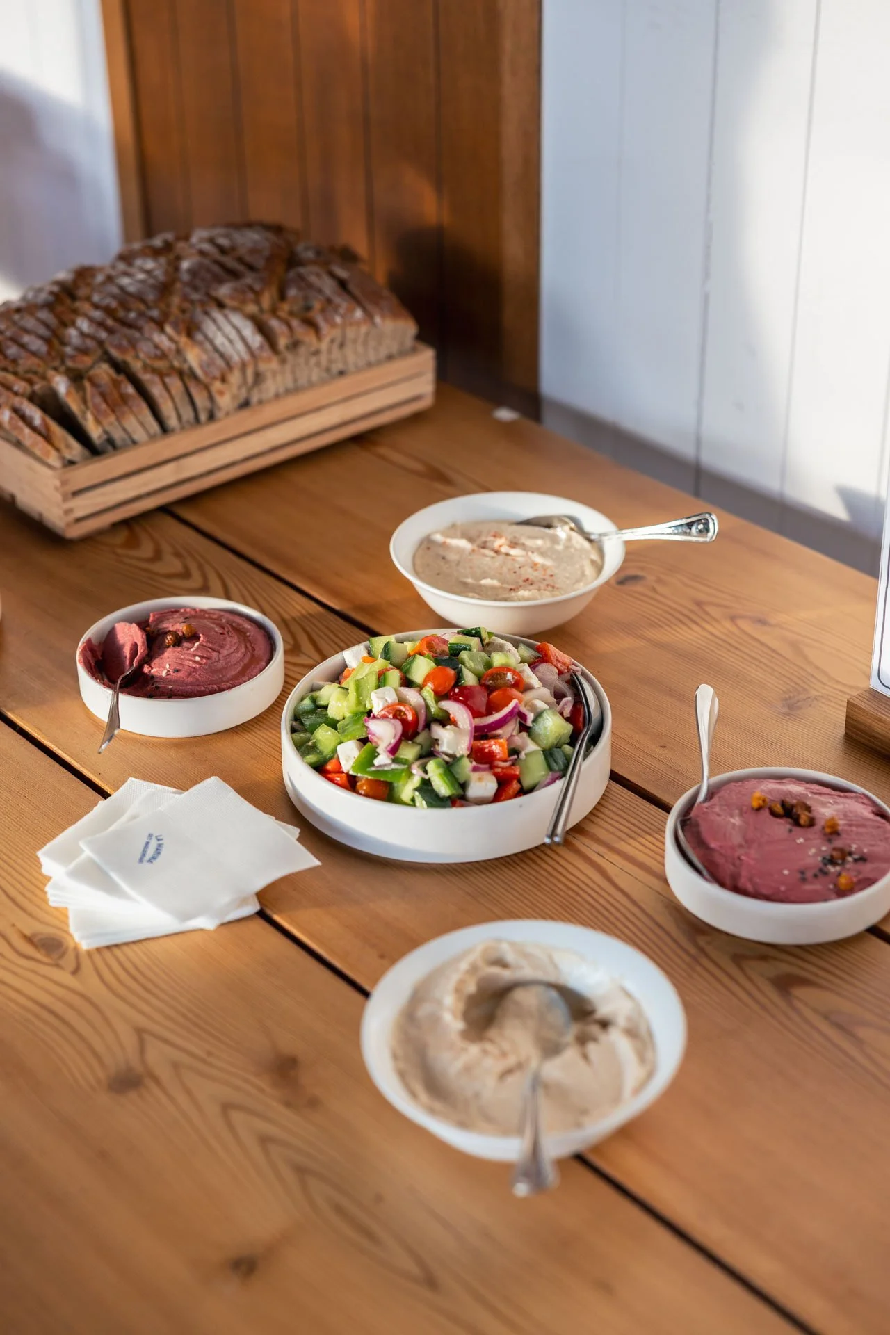 Assortiment de salades et de dips sur une table en bois, avec un pain en tranches dans une caisse en bois, dans un cadre intérieur.