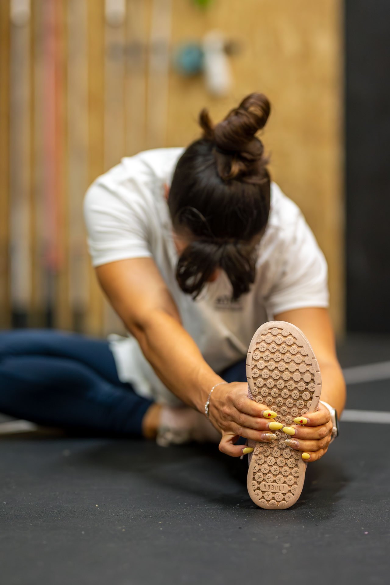 Une femme fait des étirements au sol, portant un t-shirt blanc, des pantalons foncés, et tenant une chaussure en caoutchouc beige avec des motifs. La femme a les cheveux attachés en un chignon et a les ongles peints en jaune.