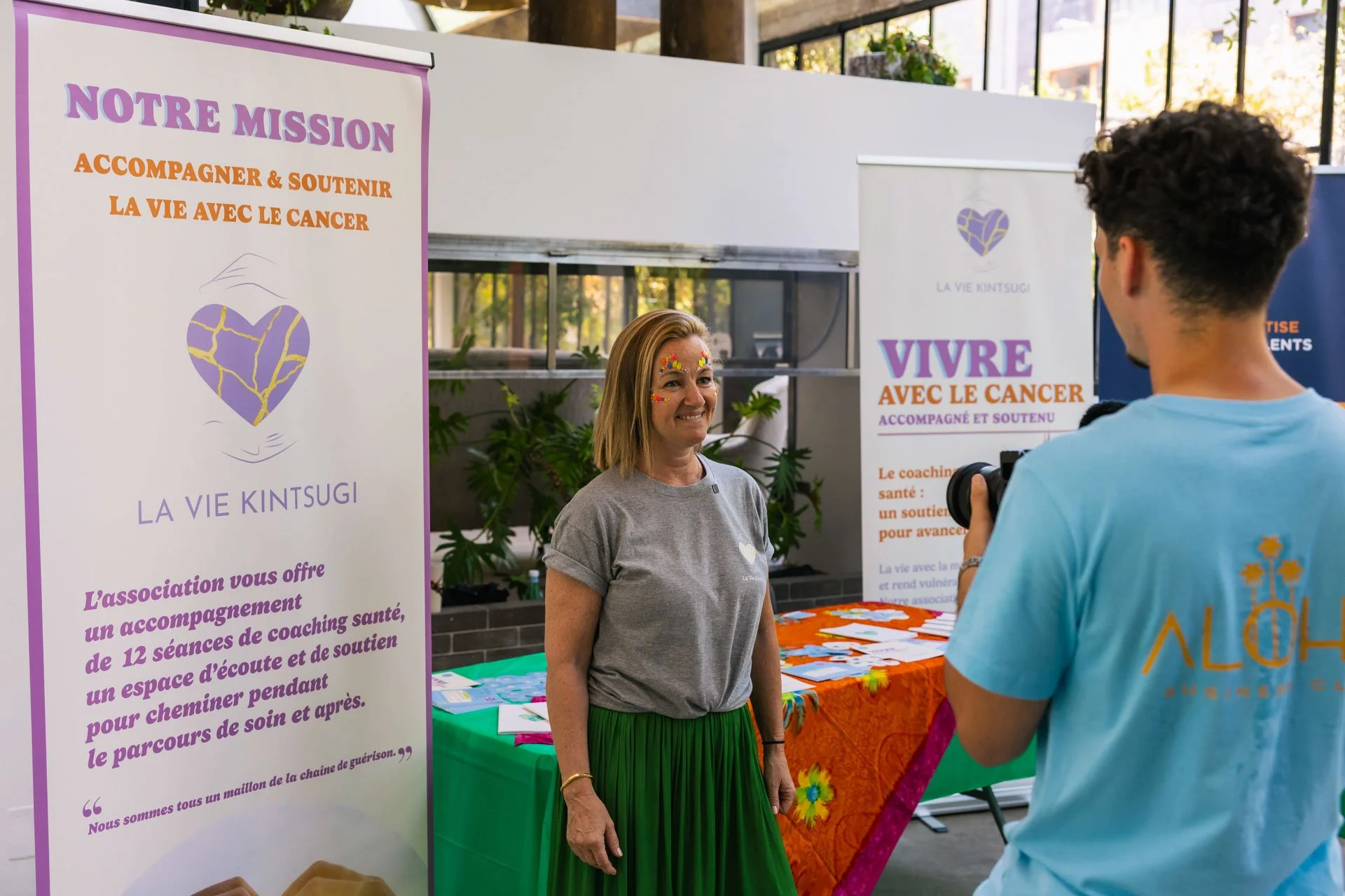 Une femme parle avec un homme dans un stand d'association. L'affiche à gauche indique la mission de l'association: accompagner et soutenir la vie avec le cancer, avec des textes en français et un logo en forme de cœur violet et or. Le stand a un fond