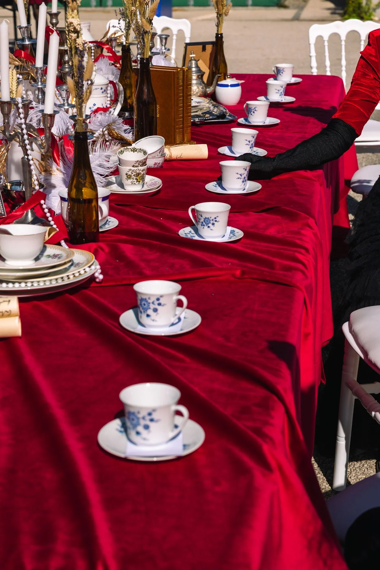 Une table dressée avec une nappe rouge, des tasses à café en porcelaine blanche ornées de motifs bleus, des assiettes, des chandeliers en argent, des bouteilles en verre foncé, et diverses décorations, suggérant une mise en place pour un repas ou une