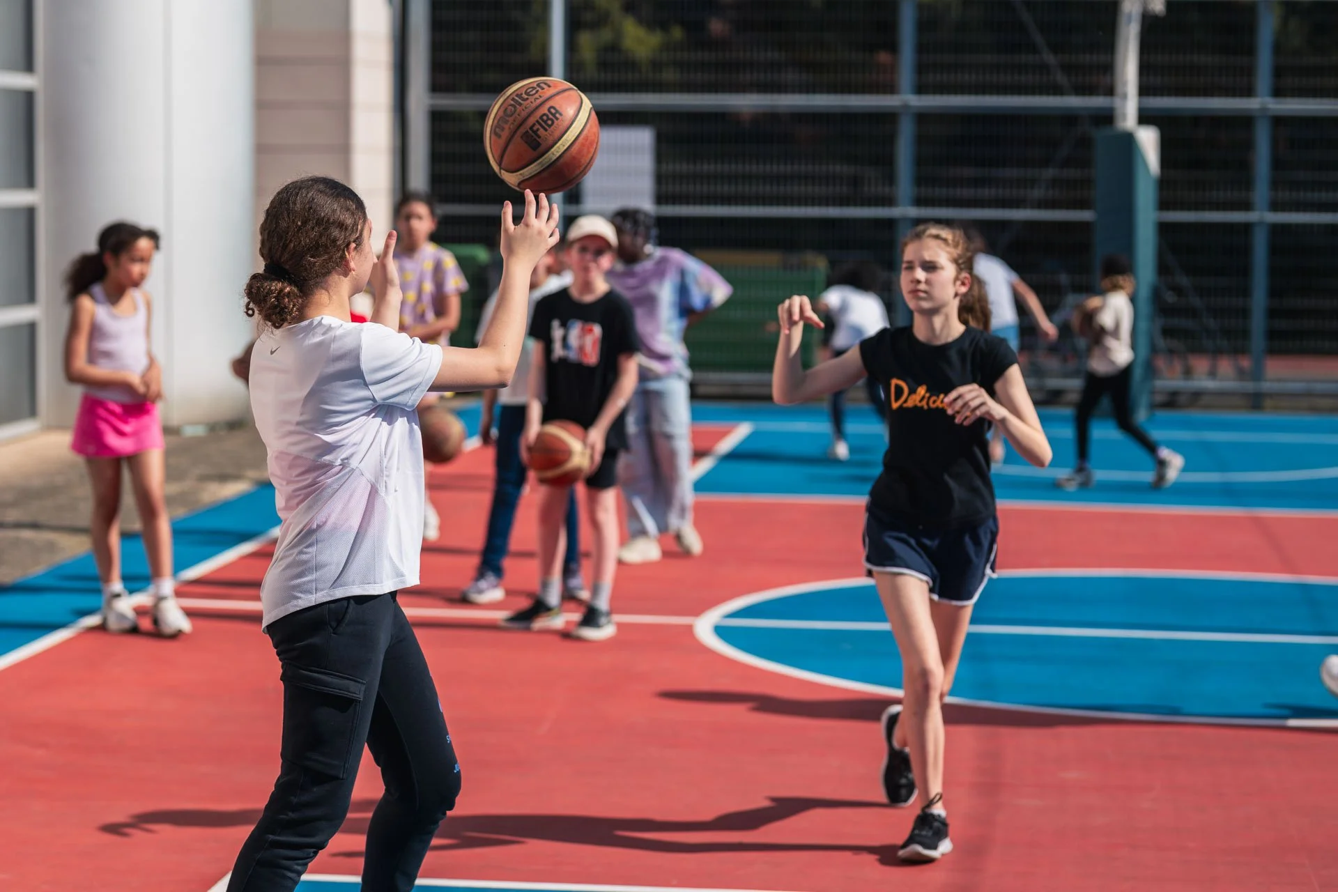 Des jeunes jouent au basketball sur un terrain extérieur. La fille en avant tire un ballon dans la direction d'une autre fille, tandis que d'autres enfants se tiennent ou se déplacent en arrière.
