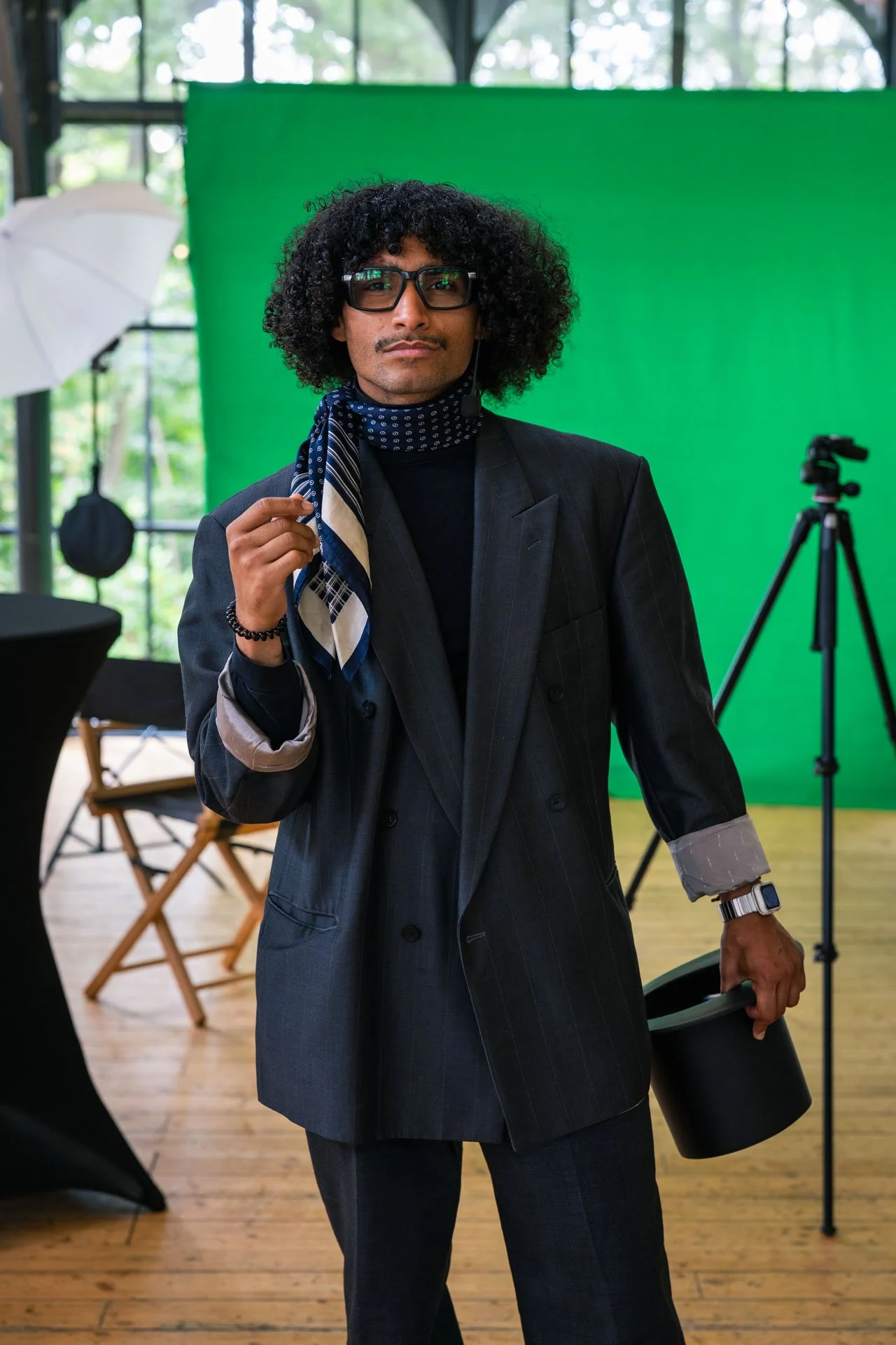 Homme portant un costume noir avec une écharpe à pois bleus, lunettes et un casque, posant dans un studio avec un fond vert et un équipement de tournage.