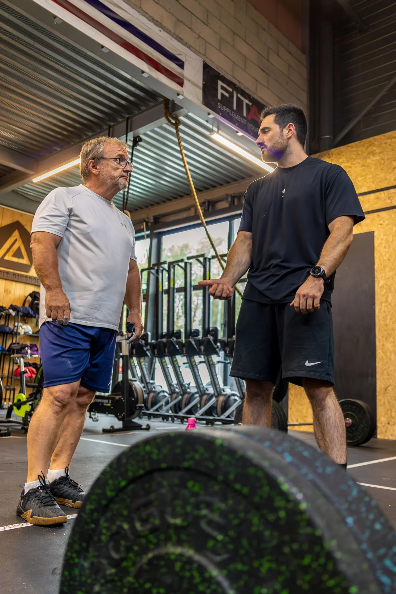 Deux hommes discutent dans une salle de sport, avec des poids et des équipements d'entraînement en arrière-plan.