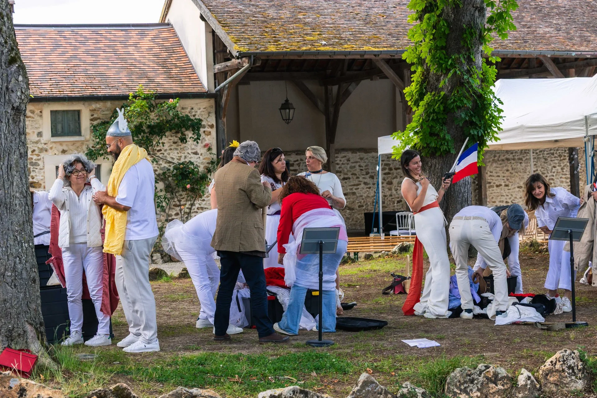 Groupe de personnes habillées de blanc, certaines portant des costumes ou accessoires, participant à une activité en plein air avec un drapeau français, entourage d'arbres et de bâtiments en pierre.