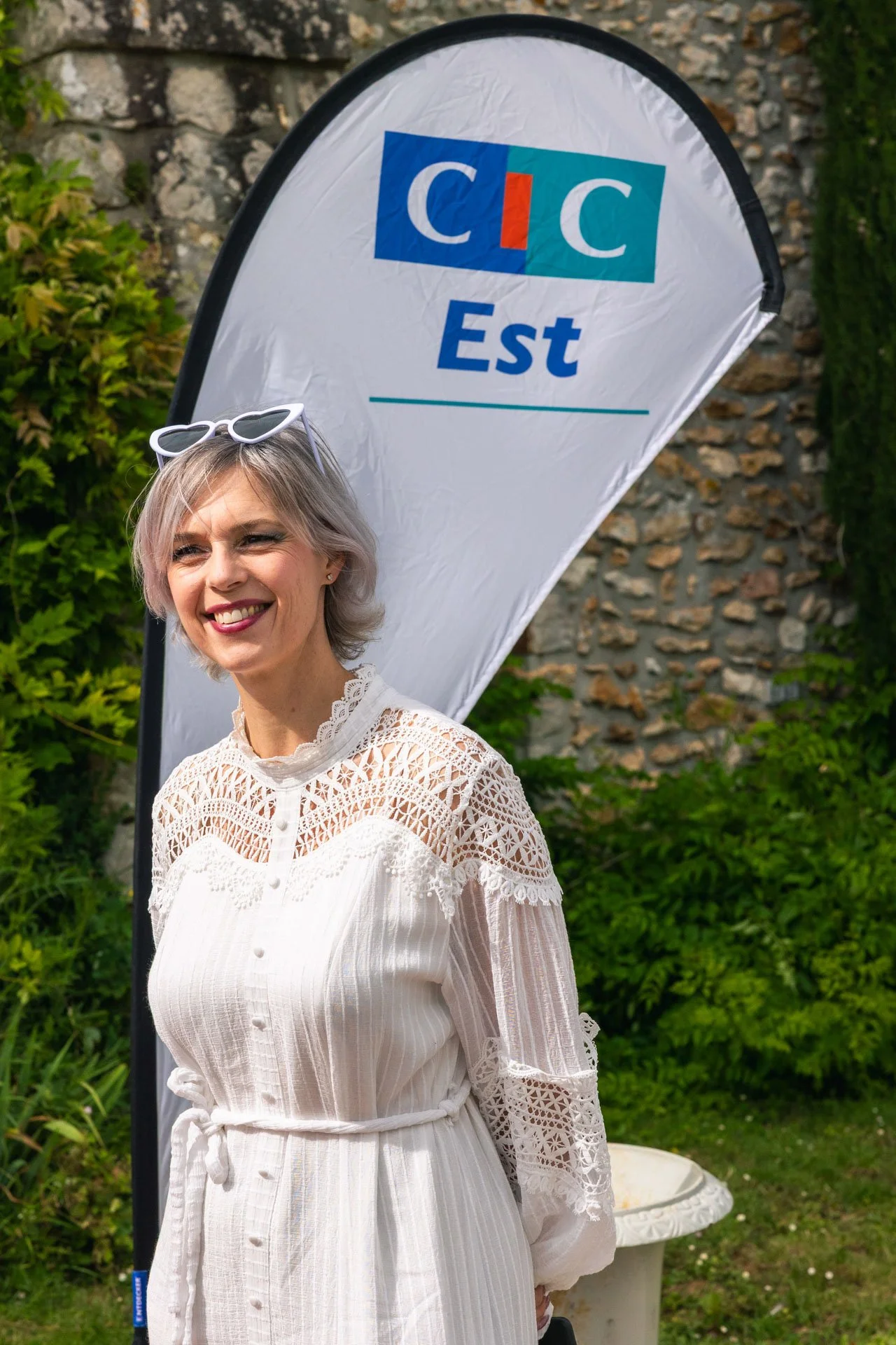 Une femme souriante portant une robe blanche avec des détails en dentelle, portant des lunettes de soleil blanche sur la tête, se tenant près d'un drapeau avec le texte 'CIC Est', en extérieur avec des plantes vertes et un mur en pierre en arrière-pl