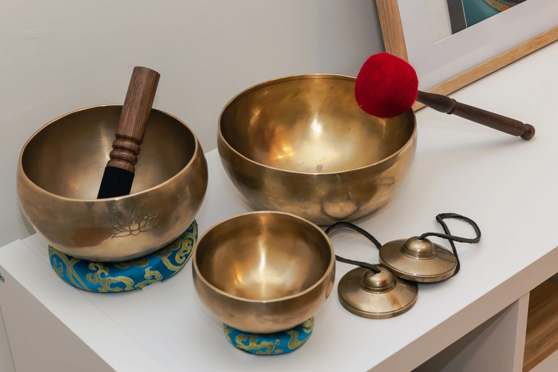 Ensemble de bols chantants tibétains de différentes tailles, certains avec de la décoration, placés sur une table blanche. Deux tambours en bois et une cloche en métal avec un cordon sont aussi visibles.