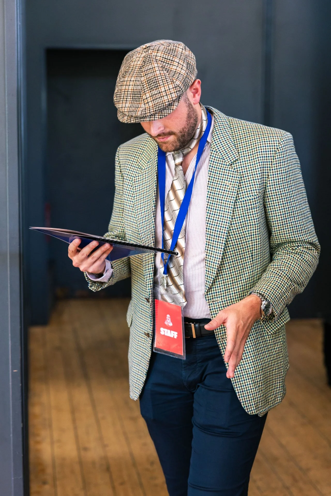 Un homme avec une barbe portant un chapeau à carreaux, une veste à carreaux, une chemise rayée, une cravate, une montre, et un badge d'équipe, regarde un document dans sa main en marchant dans un espace intérieur.