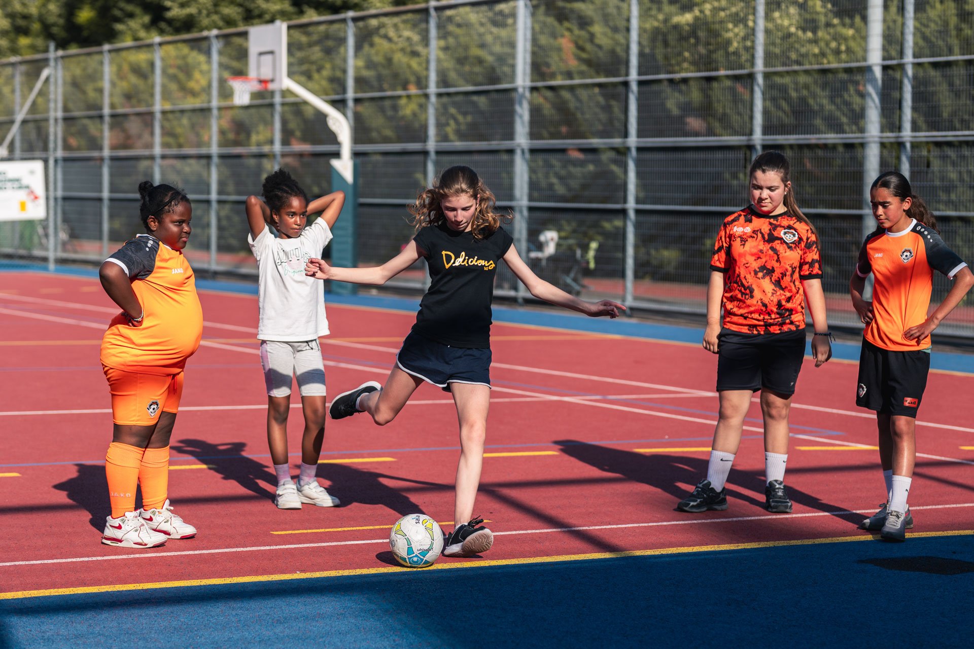 Groupe de jeunes filles jouant au football sur un terrain extérieur, avec un filet et des arbres en arrière-plan.
