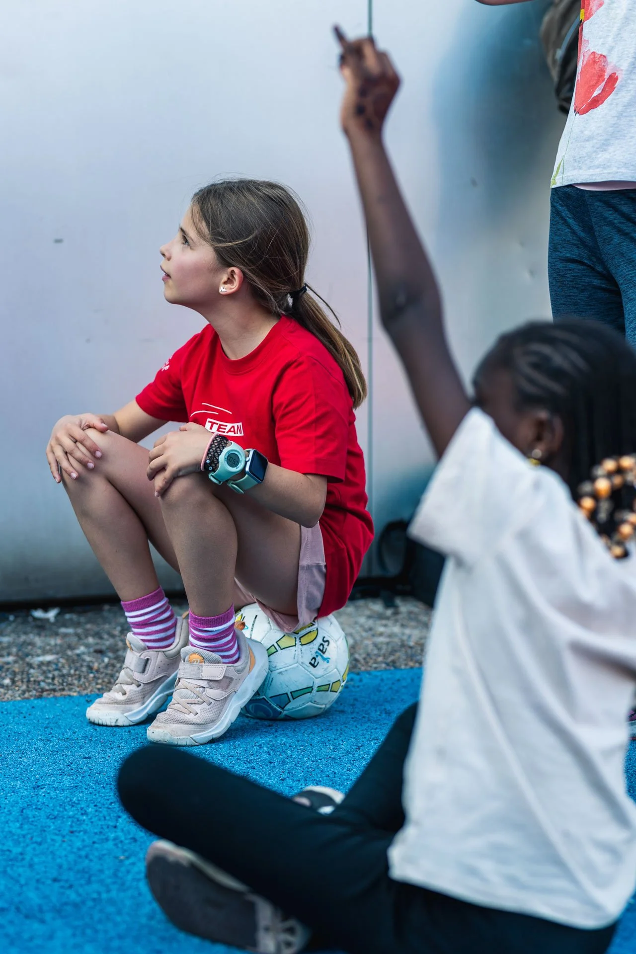 Une jeune fille assise en tailleur avec une casquette, portant une chemise rouge et des baskets, à côté d’un ballon de football, dans un environnement intérieur. Elle regarde une autre personne qui lève le bras.