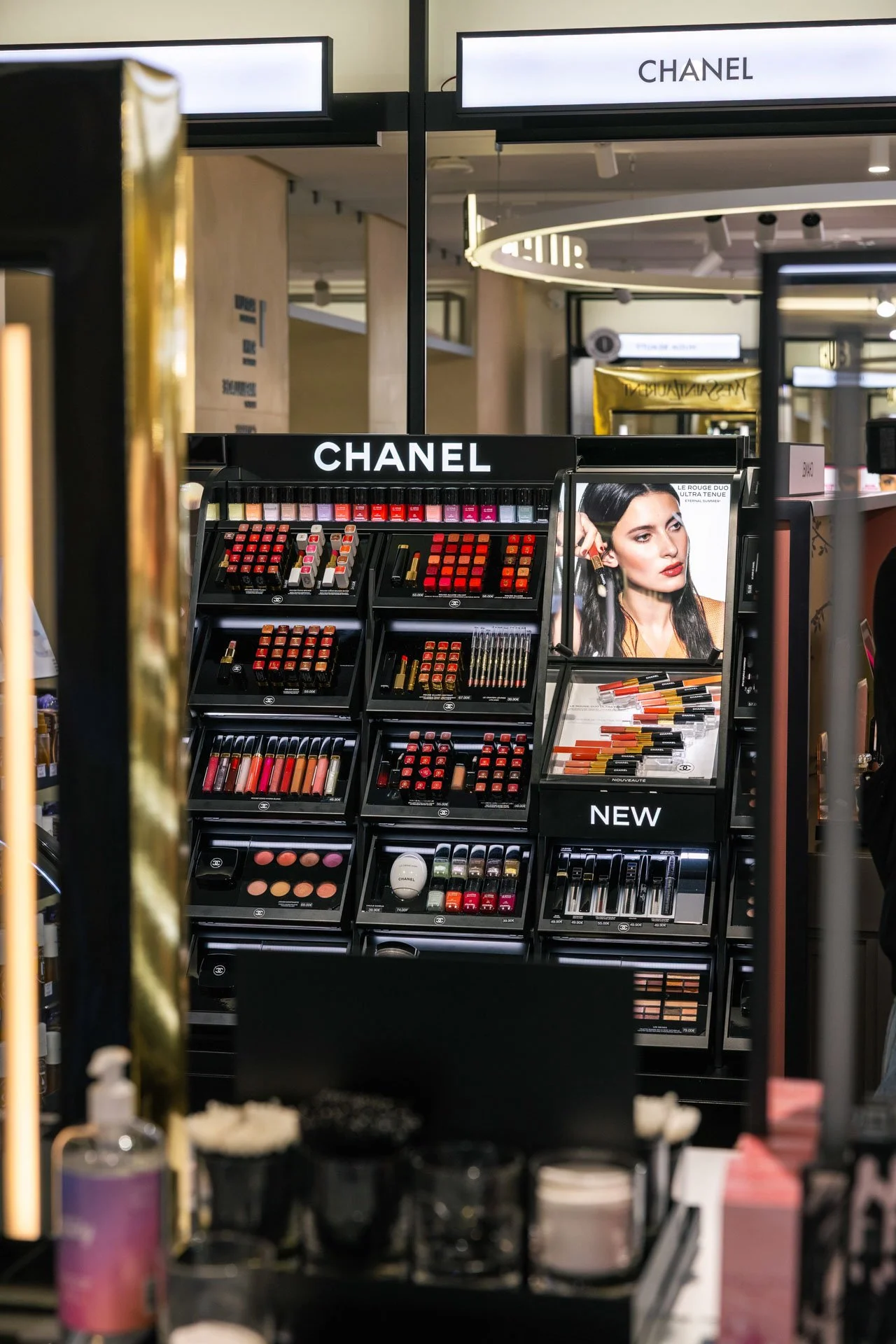 Présentoir de produits de maquillage de la marque Chanel dans un magasin, avec des palettes de rouges à lèvres, gloss, et autres cosmétiques colorés, et un panneau avec une mannequin