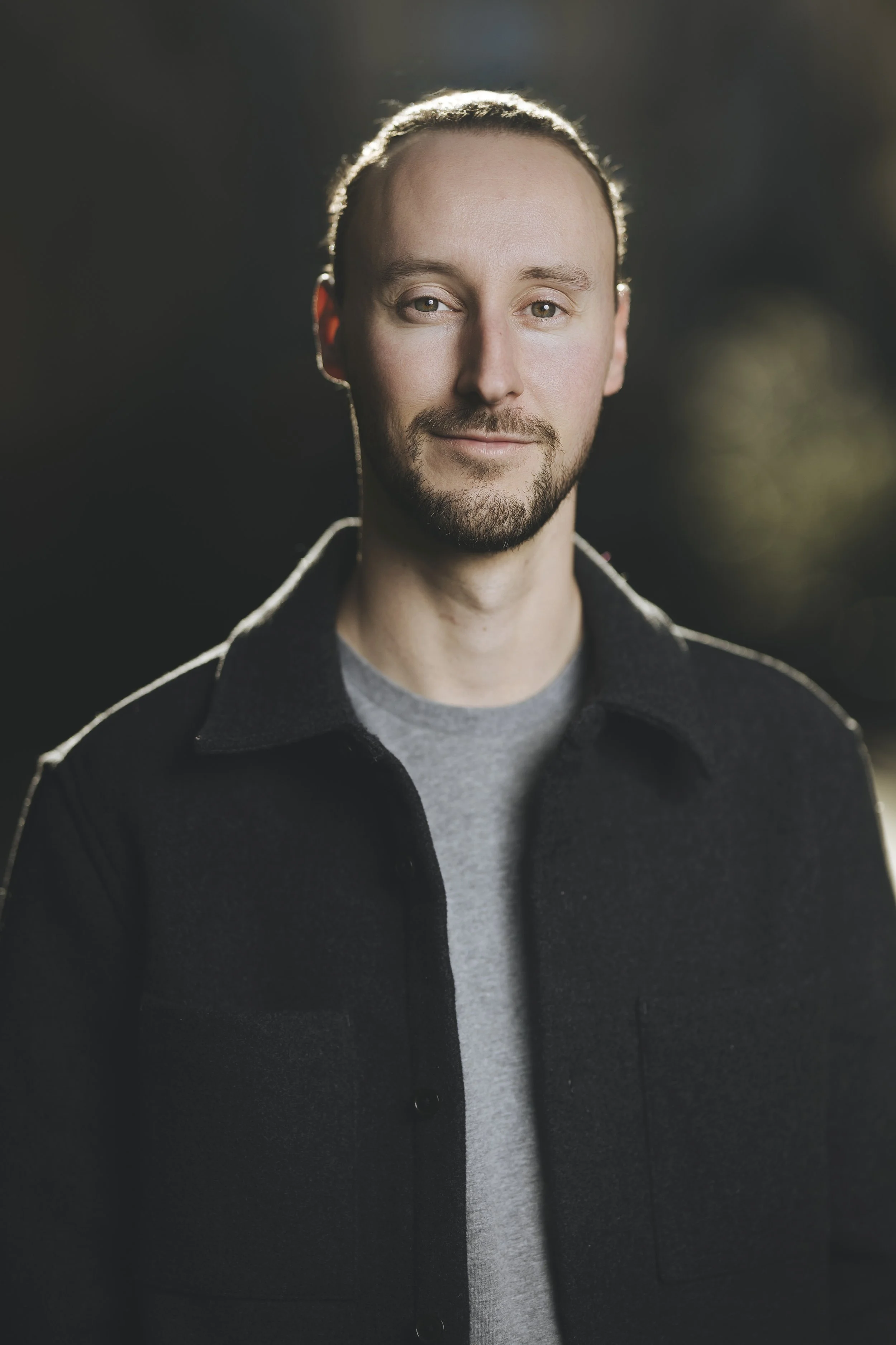 Portrait d'un homme avec barbe, portant un pull gris et une veste noire, avec un fond flou et un éclairage naturel