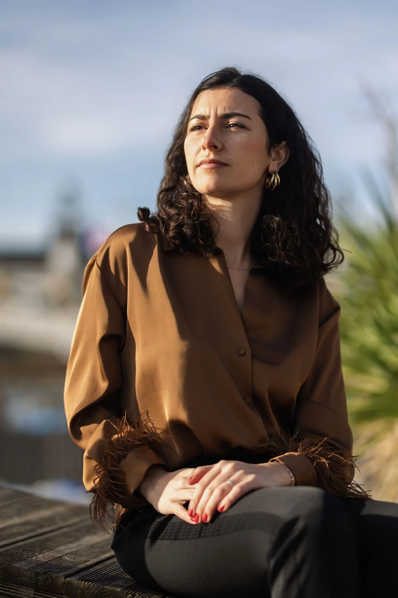 Une femme aux cheveux bouclés, portant une chemise marron avec des détails en fourrure au poignet, assise sur un banc en bois en plein air, regardant vers la droite avec un ciel bleu en arrière-plan.