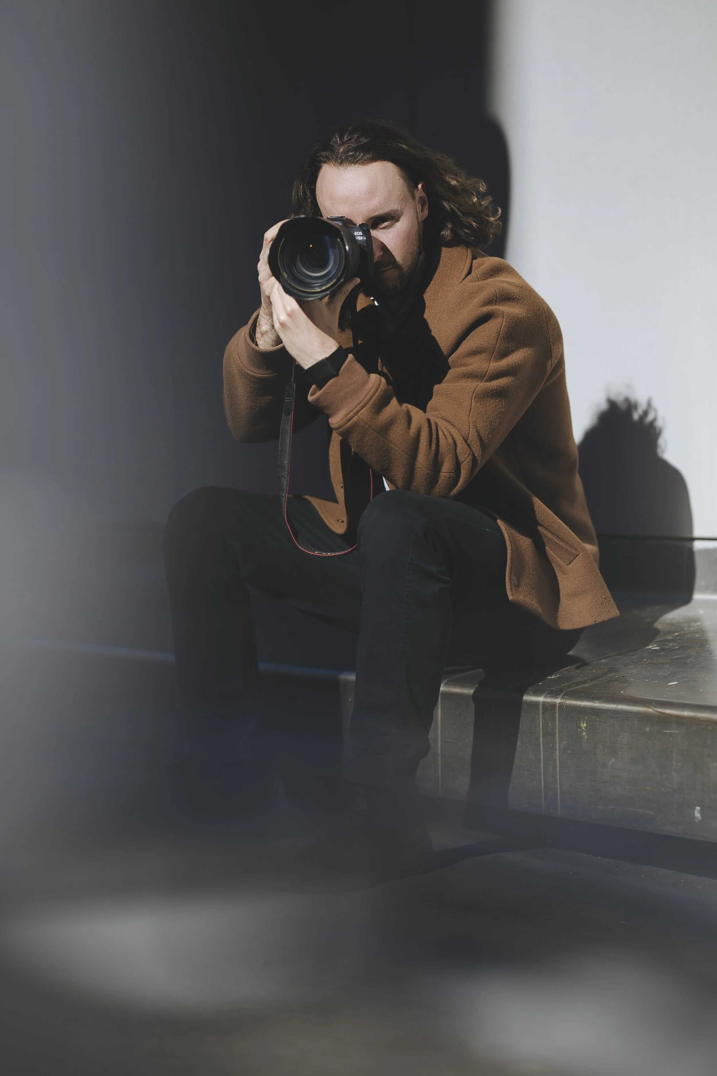 Un homme avec des cheveux longs et une barbe brune, portant une veste marron, qui prend une photo avec un appareil photo numérique. Il est assis sur un banc en métal dans un lieu intérieur ou extérieur, avec un mur gris en arrière-plan.