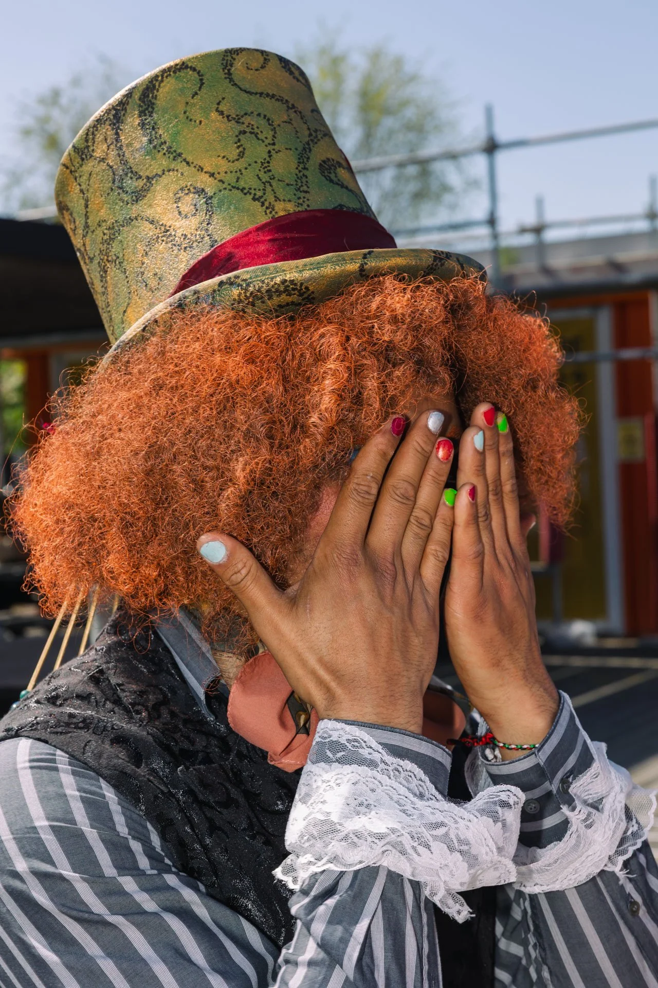 Une personne avec une perruque frisée orange, portant un chapeau haut-de-forme vert avec un ruban rouge, cache son visage avec ses mains peintes en différentes couleurs, portant un chemisier à rayures grises et blanches et un vêtement noir avec un mo