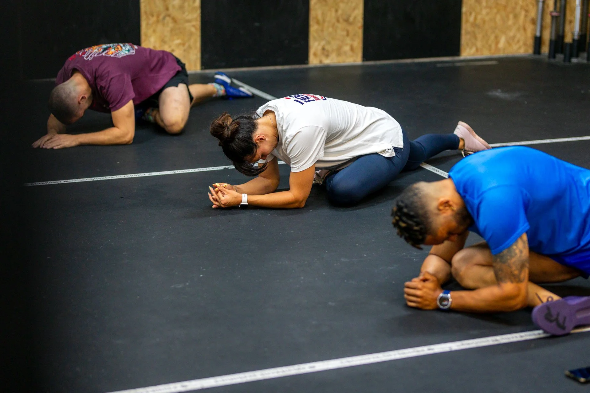 Quatre personnes en position de prière sur un sol noir dans une salle d'entraînement, la personne au centre porte un t-shirt blanc, les autres portent des t-shirts bleus ou violets.