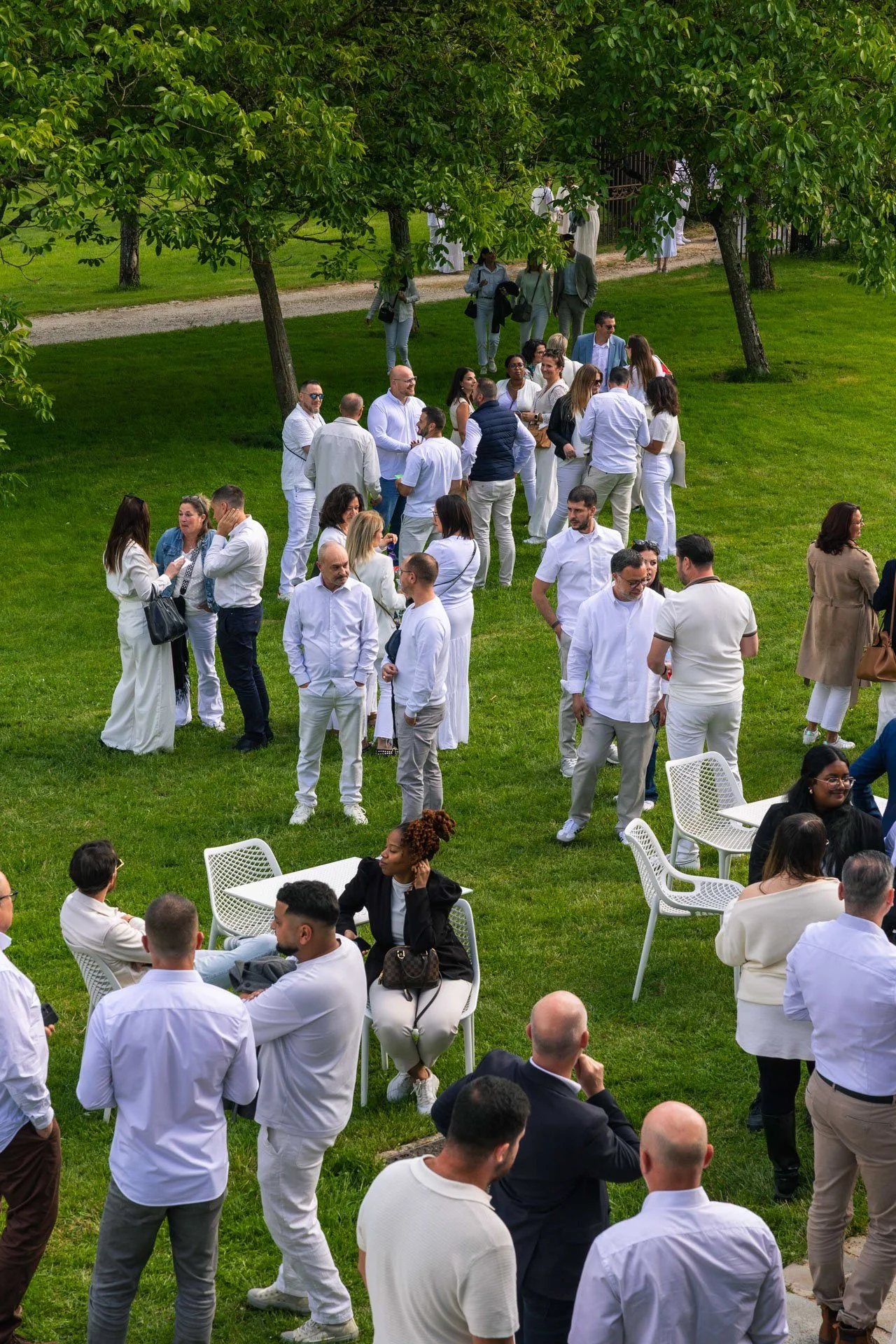 Groupe de personnes habillées en blanc, discutant et socialisant dans un espace extérieur avec des arbres et des chaises blanc.