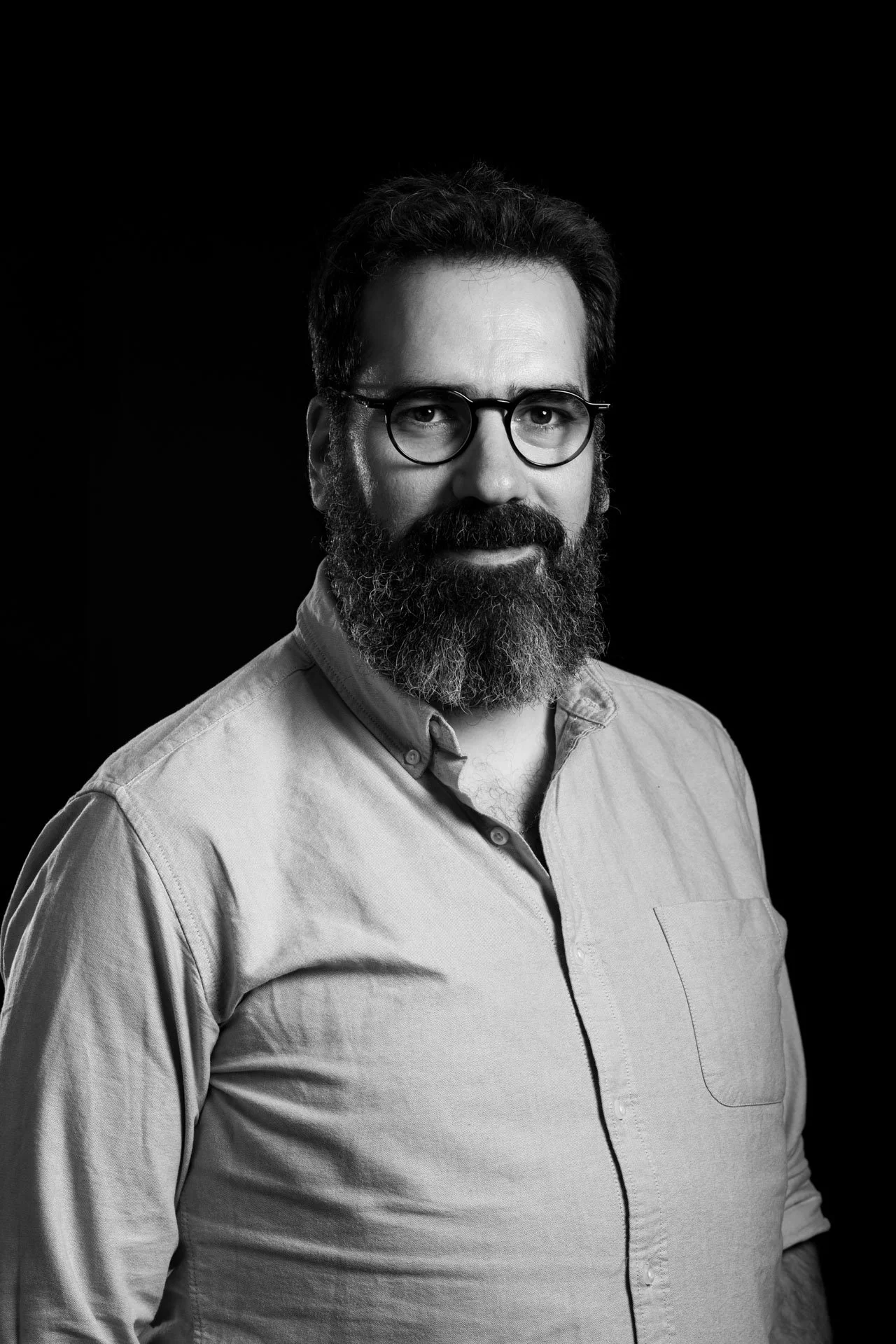 Portrait en noir et blanc d'un homme portant des lunettes, une barbe et une moustache, portant une chemise à manches longues. Fond noir.