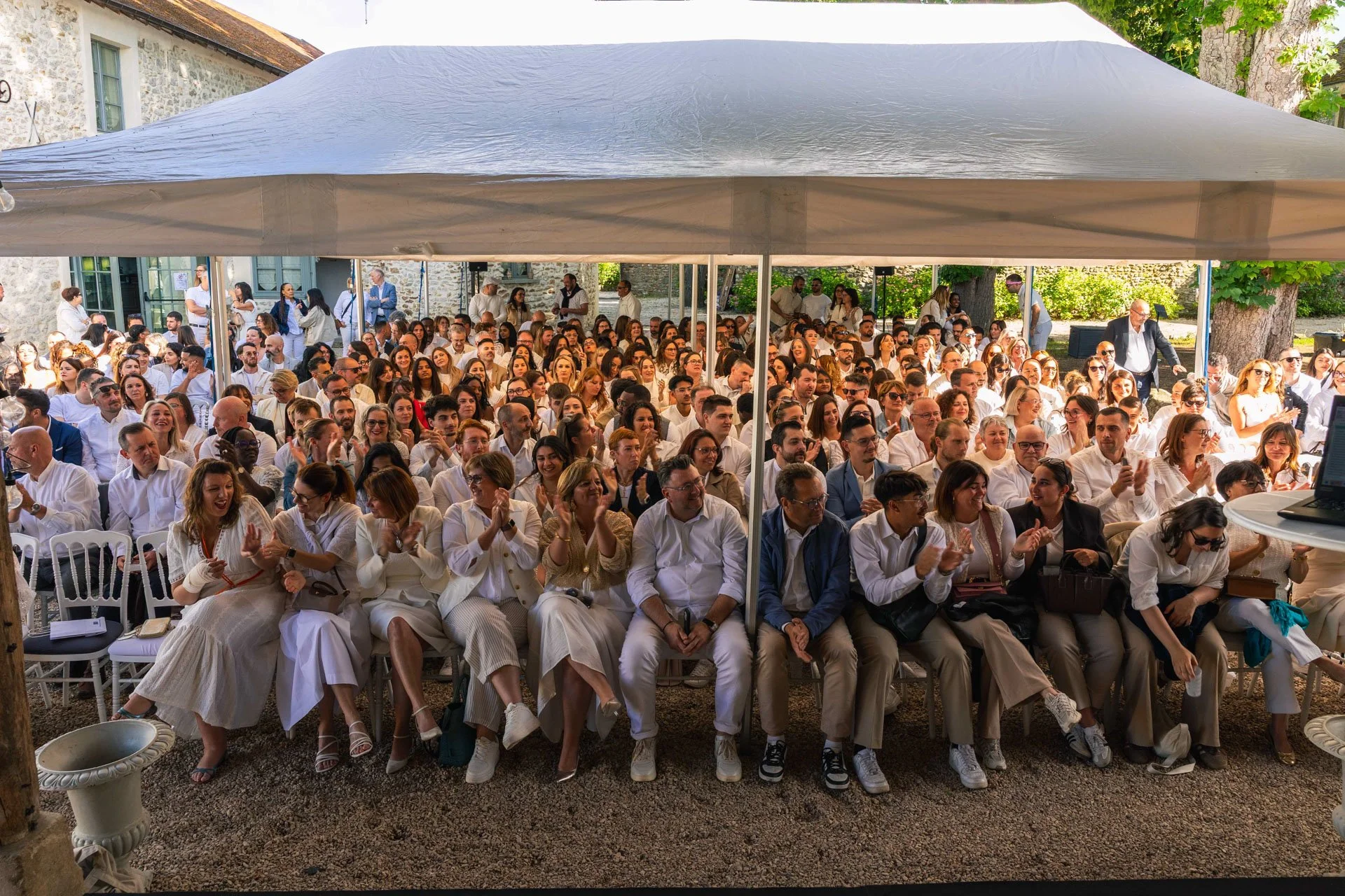 Une grande assemblée de personnes assises sous une tente lors d'un événement en plein air. La majorité portent des vêtements blancs et sourient, applaudissent ou regardent vers la scène.