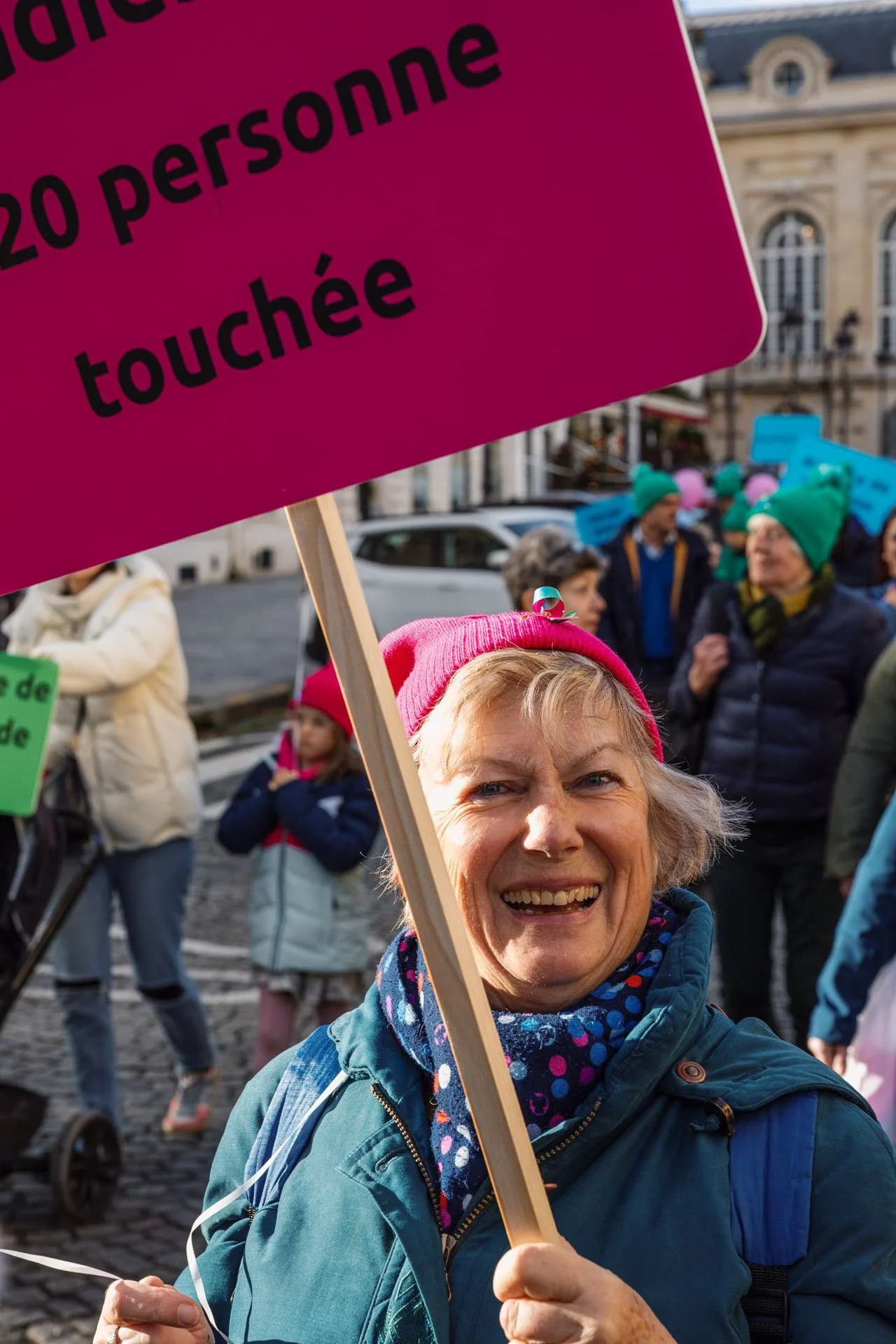 Une femme souriante tenant une pancarte rose avec du texte en français lors d'une manifestation. Elle porte un bonnet rose et un foulard à pois.