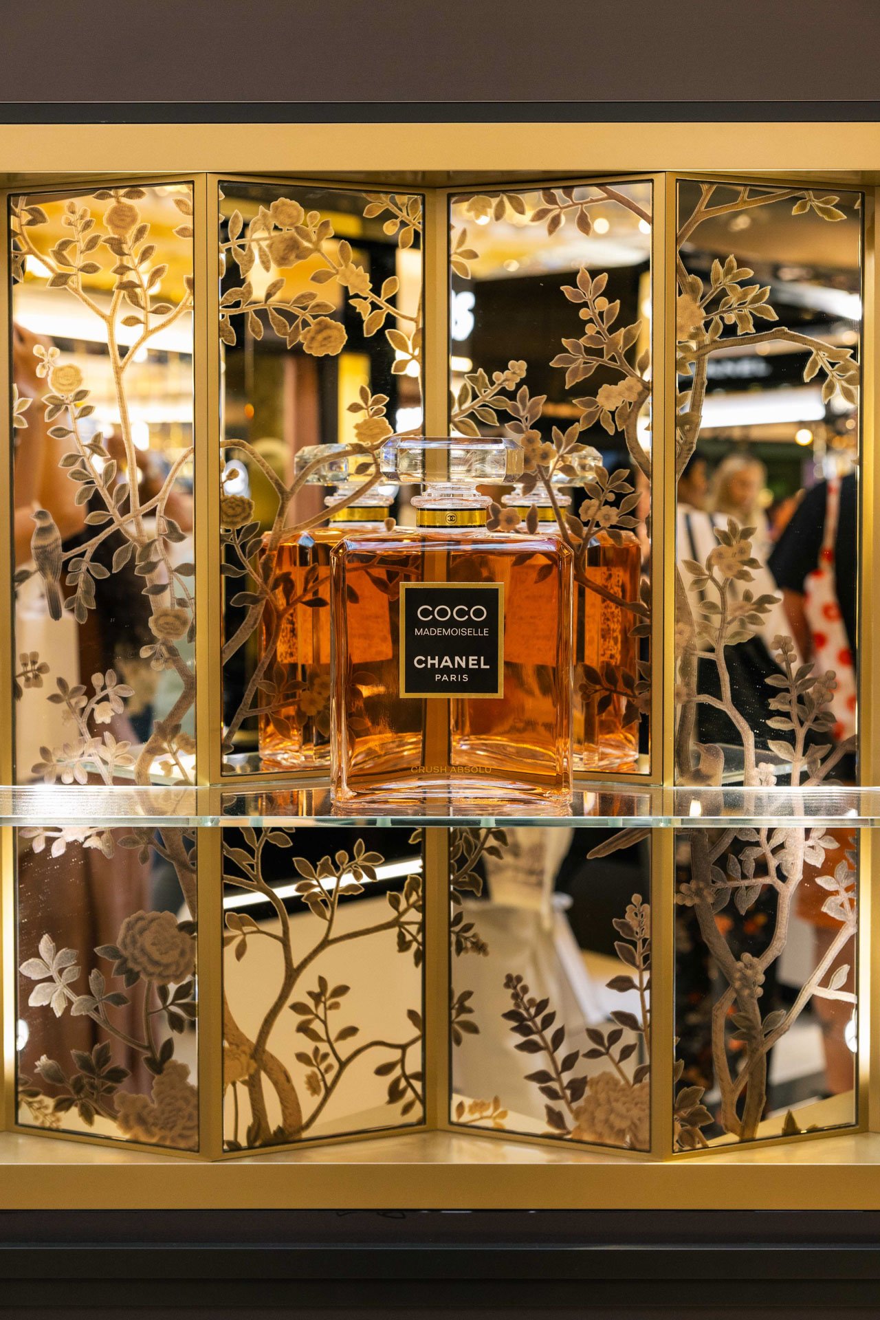 Une bouteille de parfum Chanel Coco Mademoiselle, exposée derrière une vitre décorée de motifs floraux, avec un fond flou de personnes dans une boutique ou un salon.