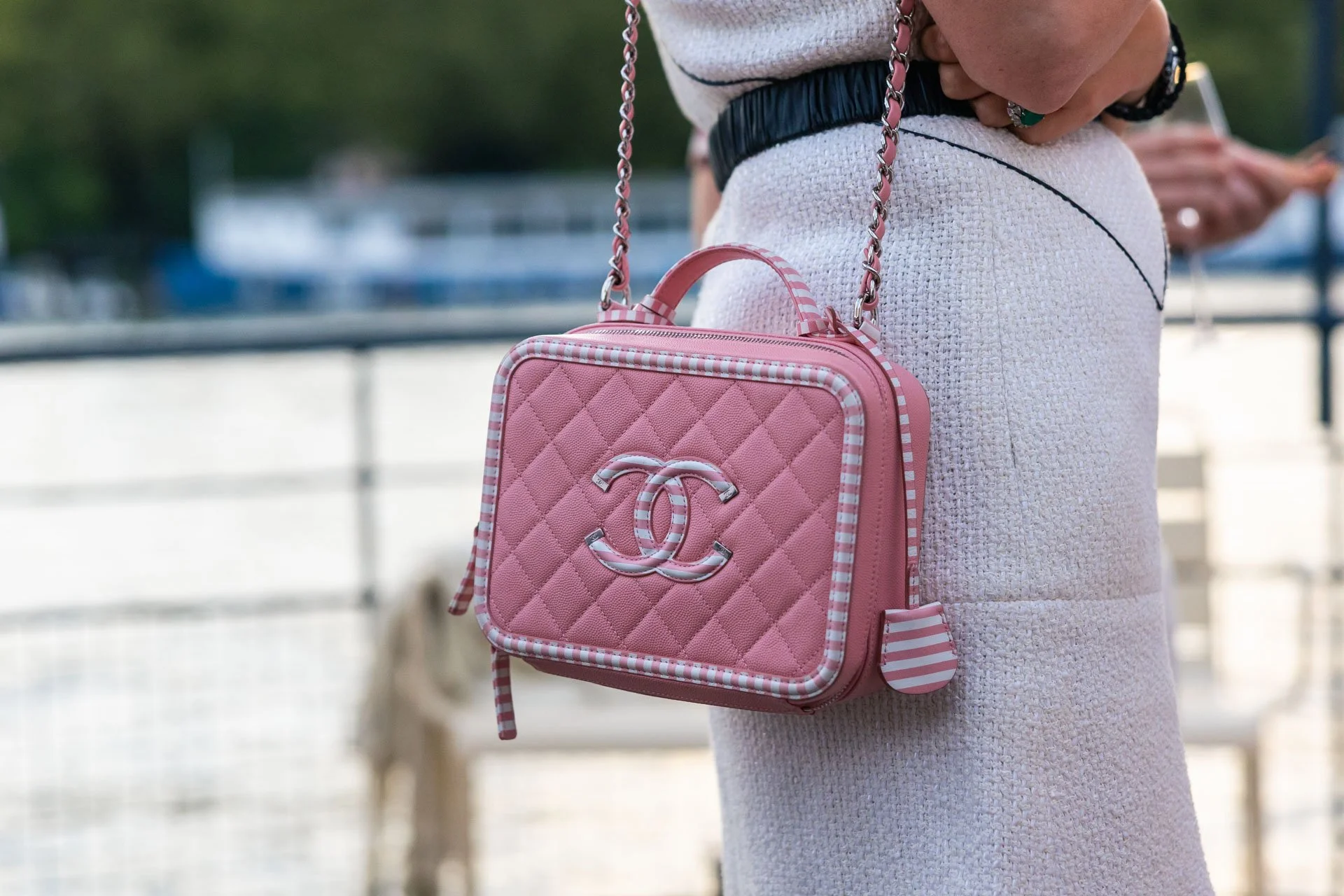 Un sac à bandoulière rose de marque Chanel porté par une personne vêtue d'un vêtement blanc avec des détails noirs, en extérieur.