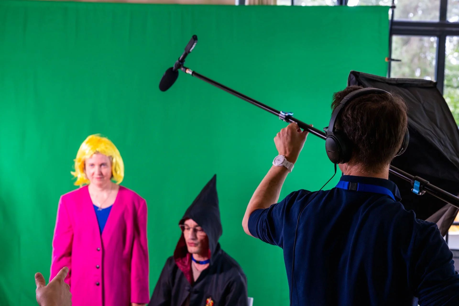 Une séance de tournage devant un fond vert avec une femme portant une wig jaune et une veste rose, et deux autres personnes portant des chapeaux de sorcière. Un technicien ajuste l'équipement de tournage.
