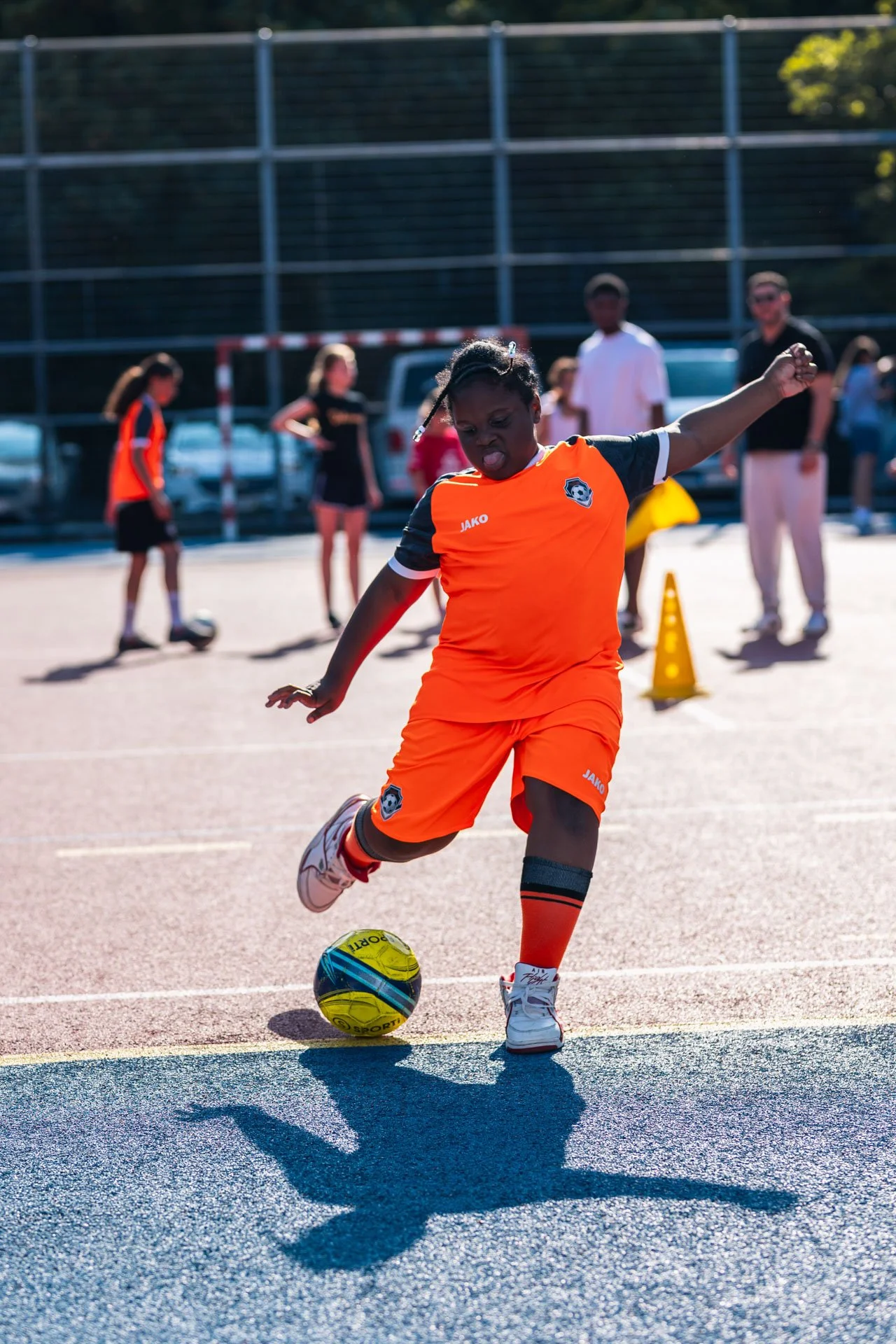 Une jeune fille joue au football sur un terrain extérieur en portant un uniforme orange. Elle est en pleine action, en train de frapper le ballon. Des personnes observent en arrière-plan, dont des enfants et quelques adultes.