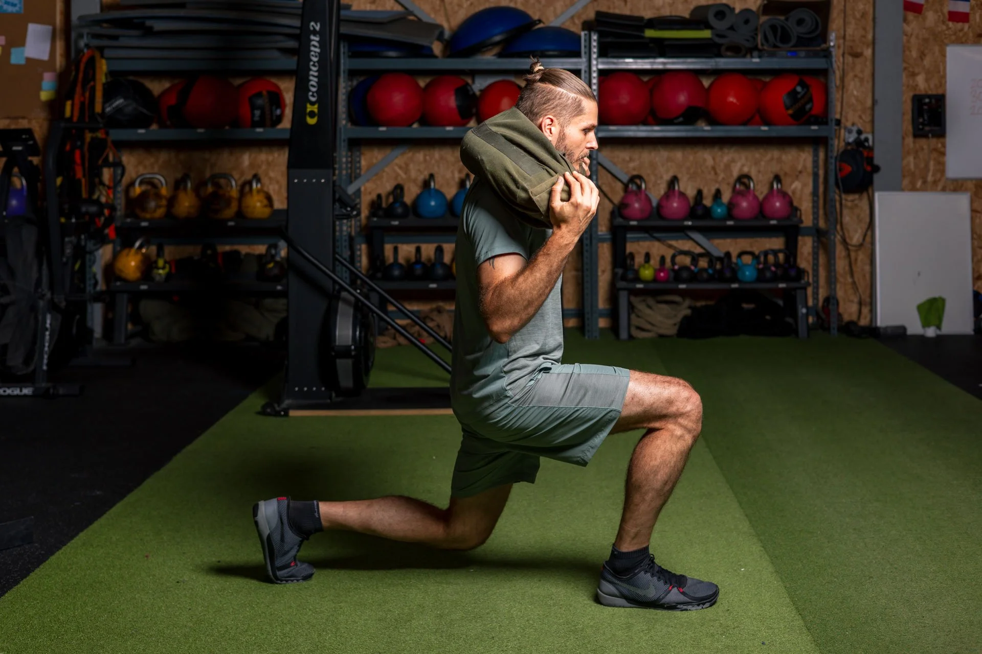 Un homme effectuant un exercice de squat dans une salle de sport, avec des poignées de kettlebell, des ballons de médecine, et des équipements d'entraînement en arrière-plan.