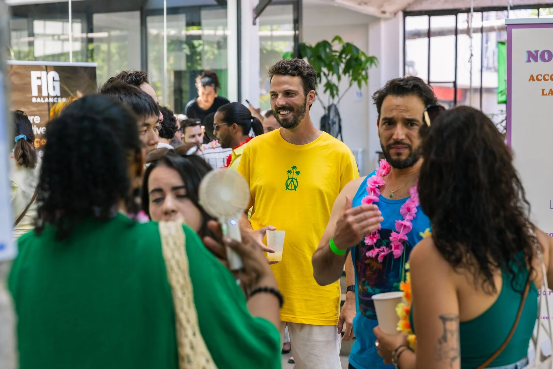 Groupe de personnes lors d'une fête ou d'un événement social avec décorations colorées, portant des vêtements colorés, dans une ambiance joyeuse à l'intérieur, avec des affiches et des plantes en arrière-plan.