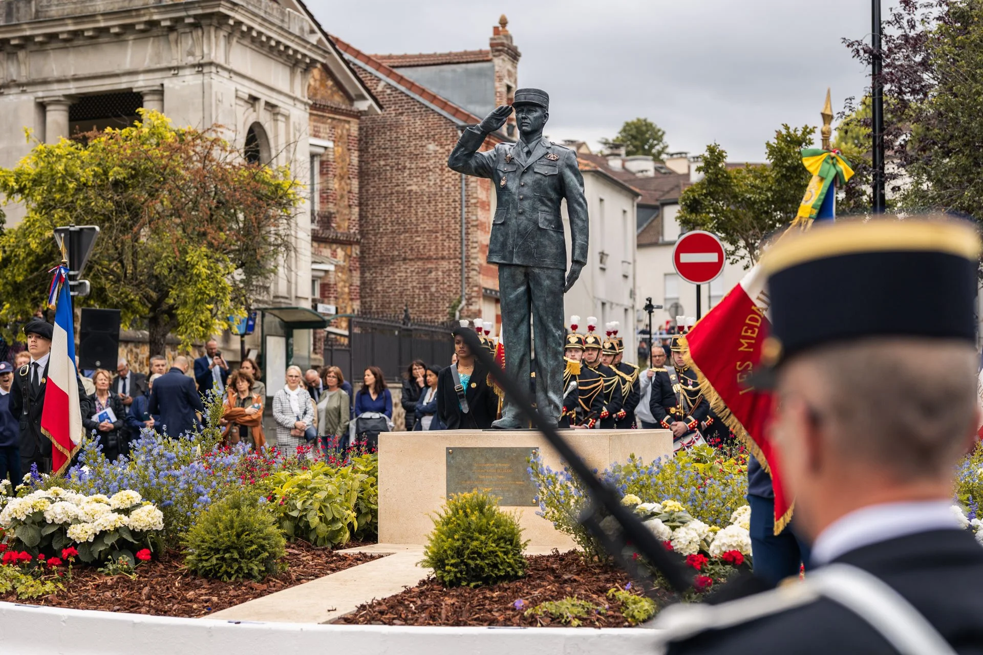 Cérémonie commémorative avec une statue en l'honneur d'une figure militaire, une foule de personnes, des soldats en uniforme, et des fleurs colorées autour de la statue, dans une ville avec bâtiments en arrière-plan.