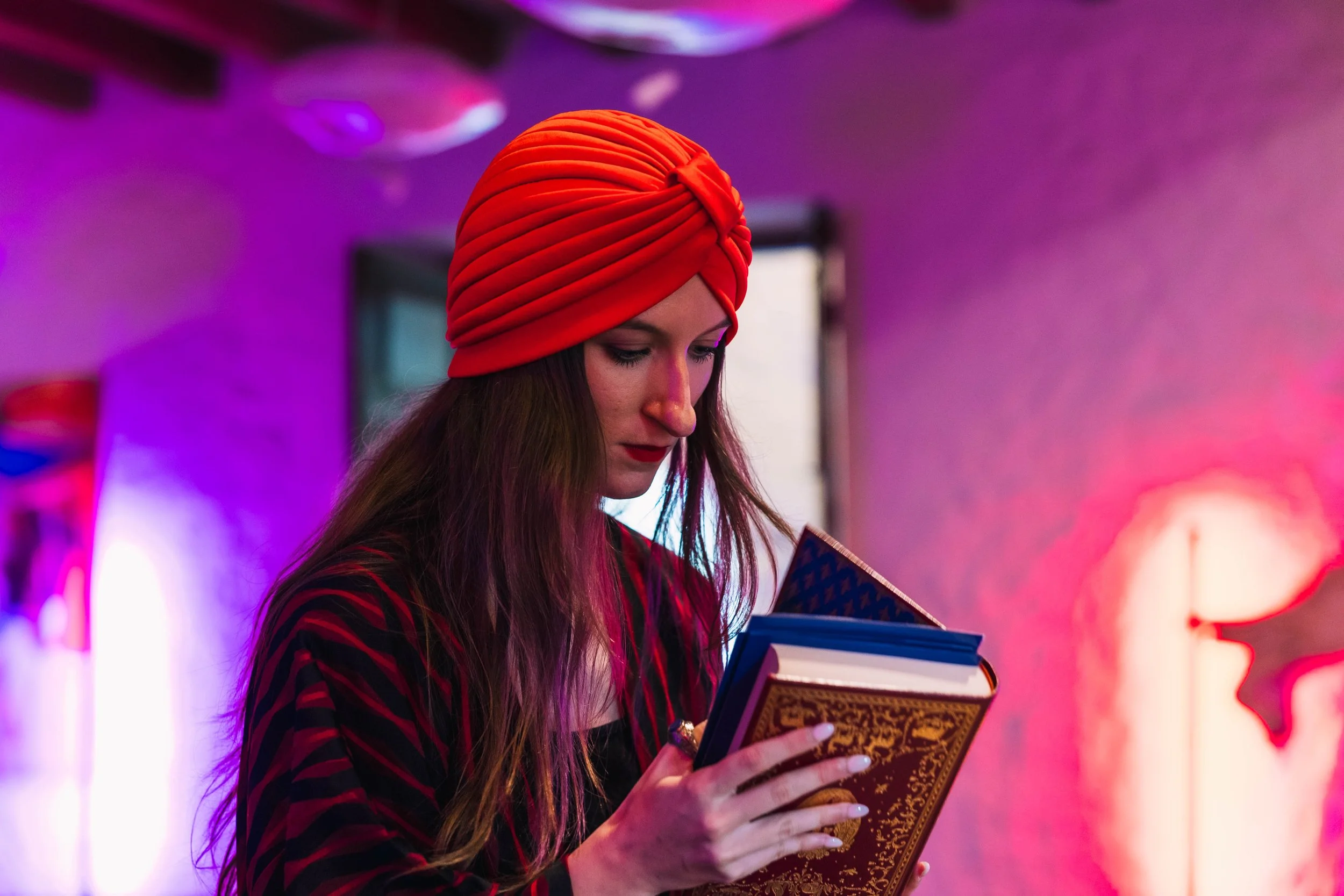 Une femme portant un turban rouge regarde un livre dans ses mains dans un environnement lumineux coloré.