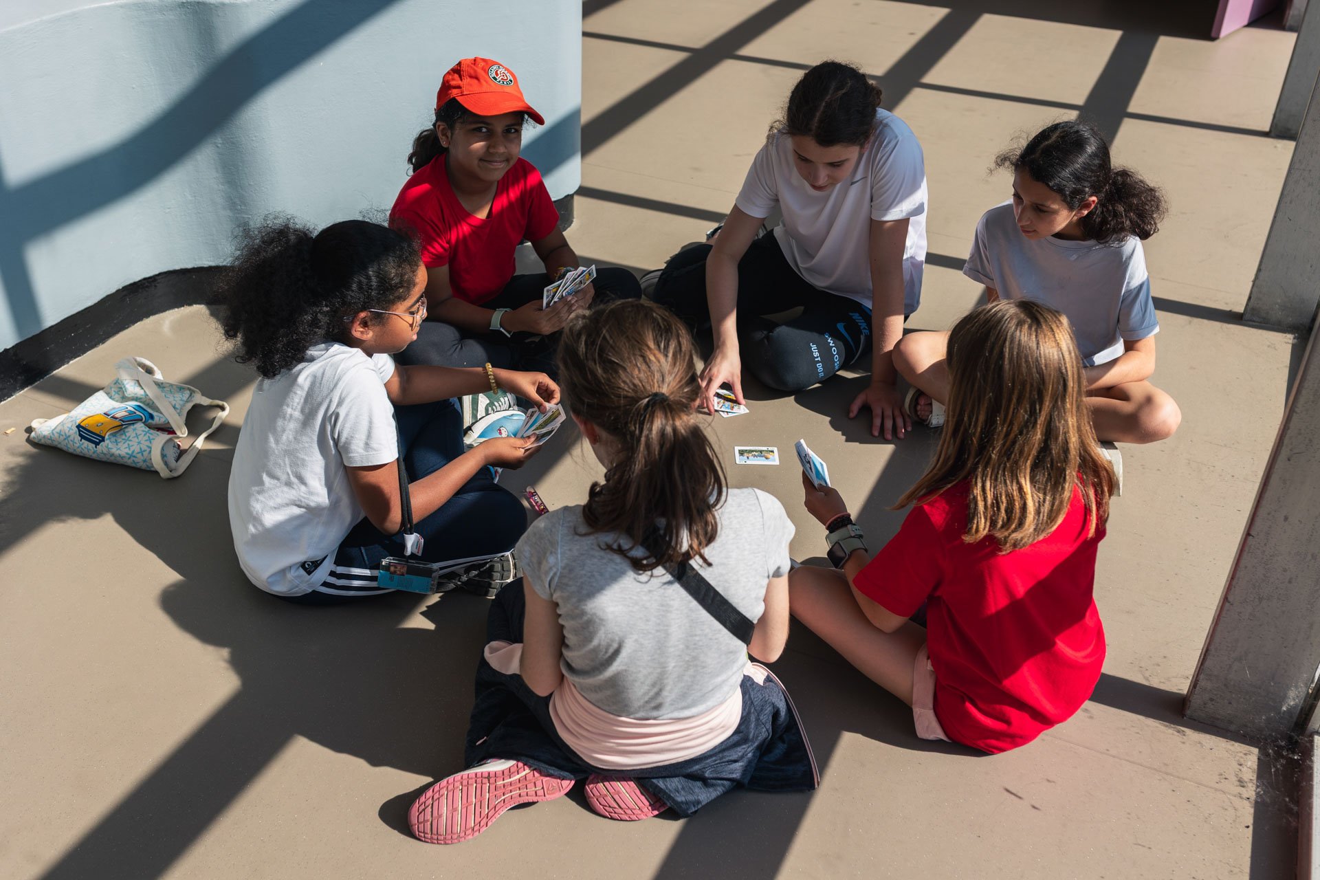 Groupe de jeunes assis par terre, jouant à un jeu de cartes dans une zone en plein soleil avec des ombres de grille sur le sol et un mur bleu en arrière-plan.