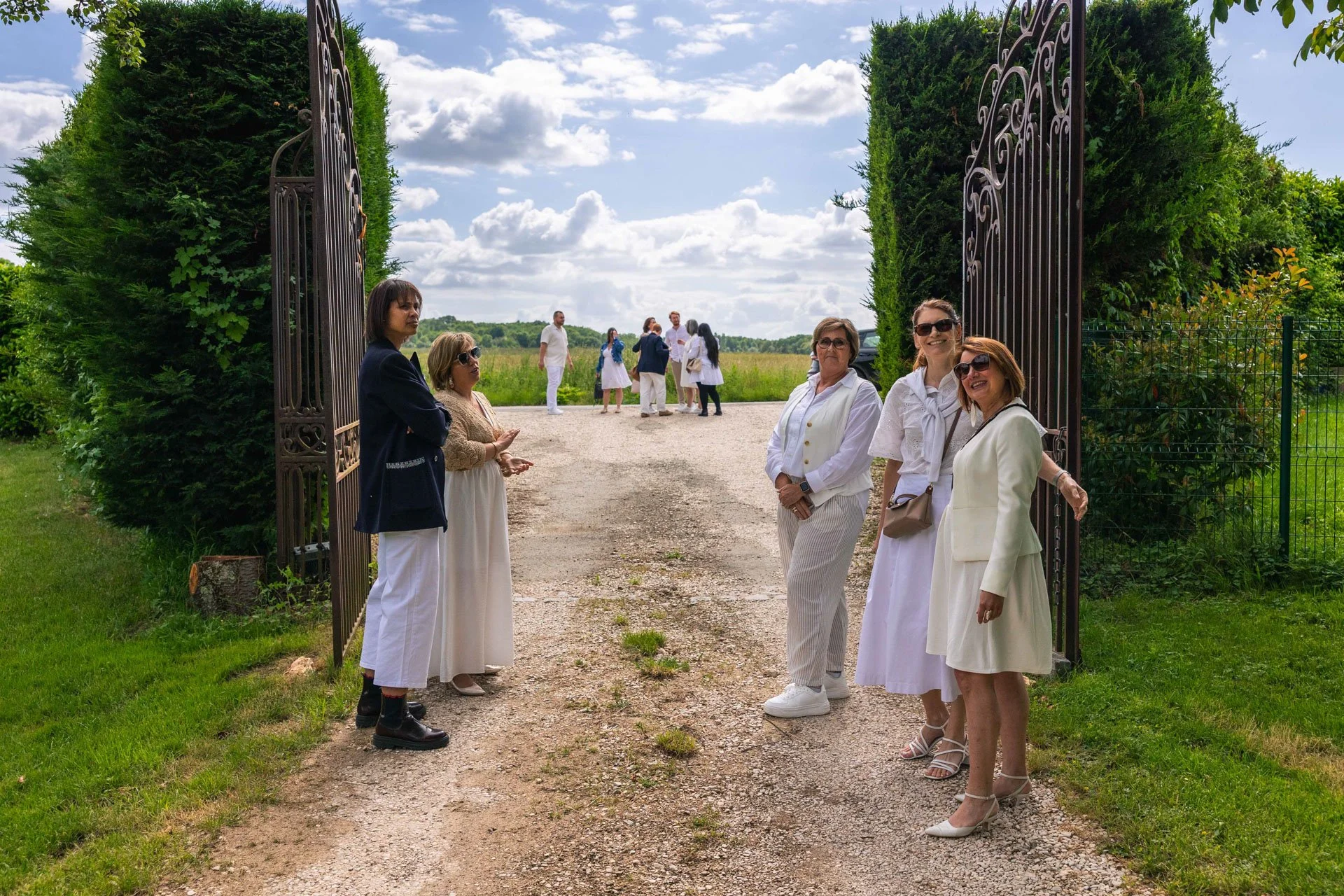 Groupe de femmes et d'hommes habillés en blanc et en vêtements décontractés, debout dans une entrée de jardin avec un portail en fer forgé et un paysage de champs et de ciel nuageux en arrière-plan.