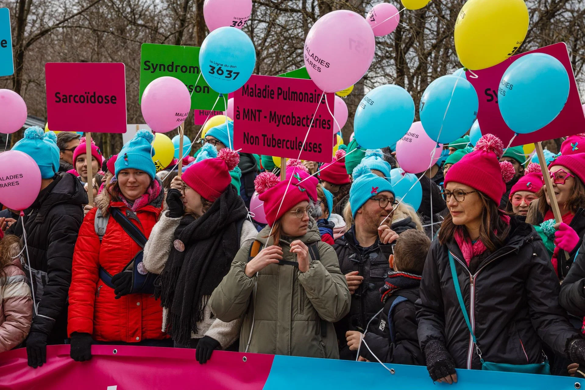 Groupe de personnes participant à une marche ou une manifestation hivernale avec des ballons colorés et des pancartes. Ils portent des vêtements chauds, y compris des bonnets et des gants, avec des messages concernant des maladies non tuberculeuses.