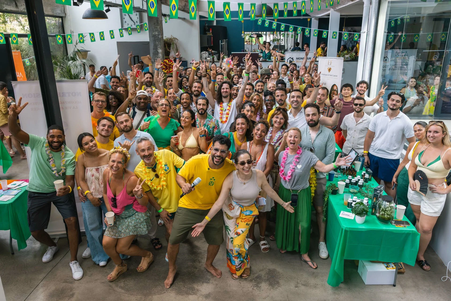 Groupe de personnes en tenue estivale, chantant, souriant, faisant des gestes de paix et de victoire, dans un décor festif avec guirlandes de drapeaux brésiliens, lors d'une célébration ou événement tropical.