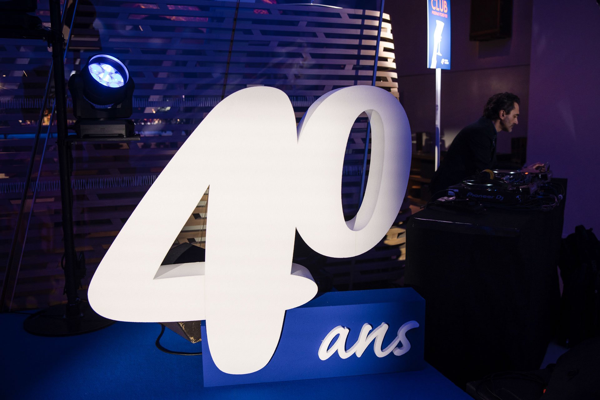 L'image montre une grande enseigne lumineuse blanche en forme de '20' sur un fond sombre, avec une personne DJ en arrière-plan, utilisant des équipements de DJ dans un environnement festif ou événementiel.